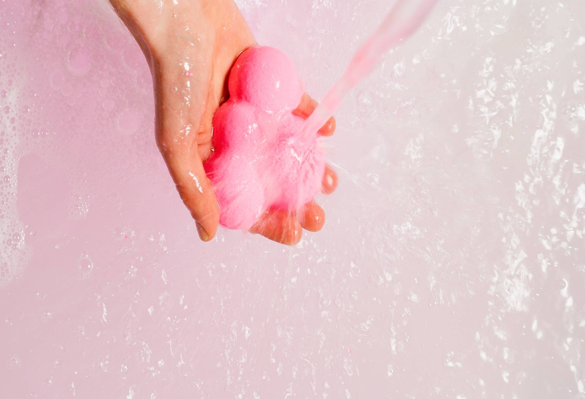 A hand holds the pink Support Bubble and water runs over it above light pink, bubbly water.