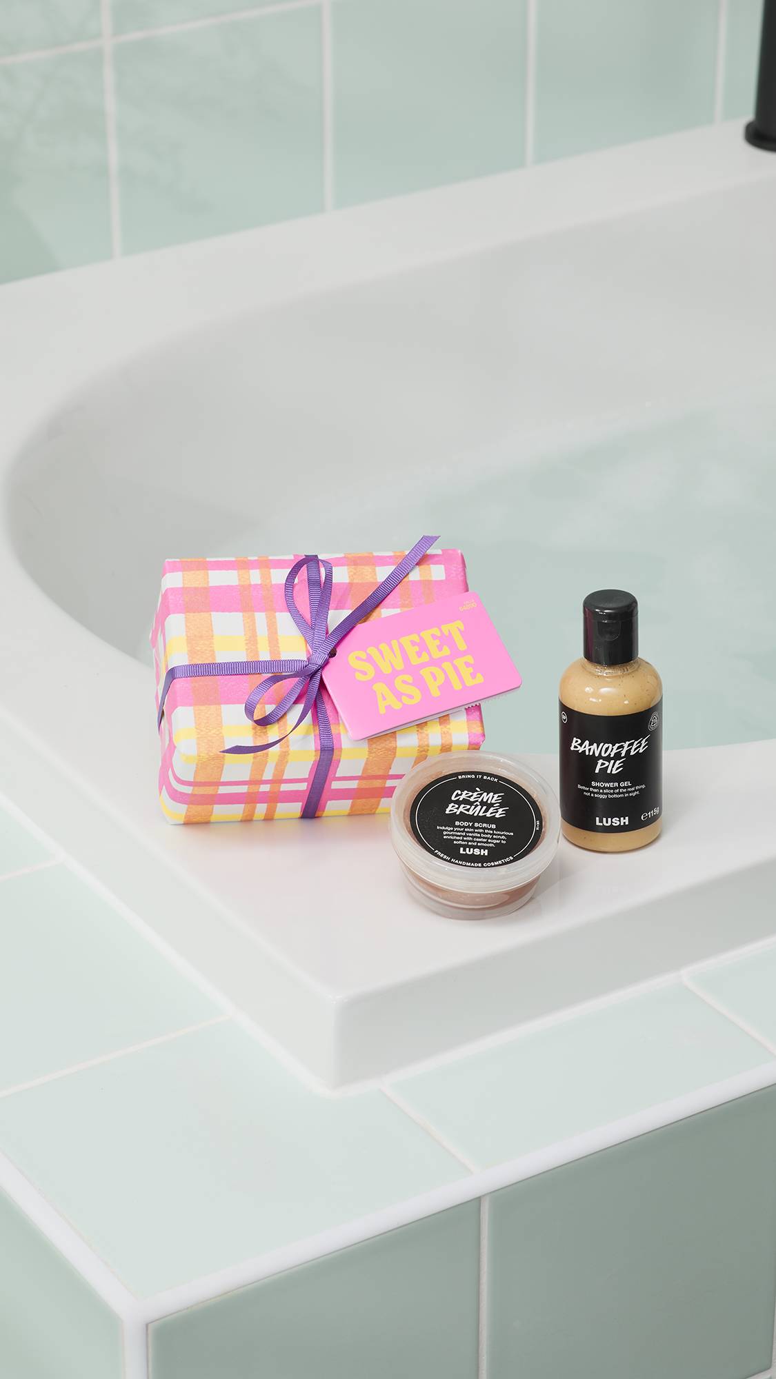 A "Sweet As Pie" gift set is displayed on the edge of a white bathtub. It includes a pink and orange checkered gift box with a purple ribbon, a pot of Crème Brûlée body scrub, and a bottle of Banoffee Pie shower gel against light green tiles.