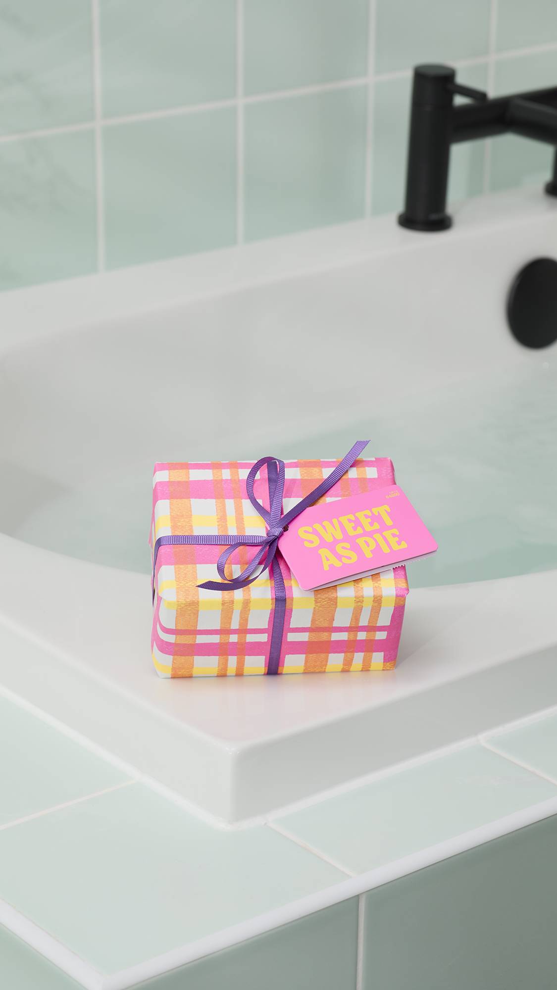 The "Sweet As Pie" gift set, wrapped in a pink and orange checkered pattern with a purple ribbon and bright pink tag, sits on the white corner of a bathtub. The background features pale green wall tiles and a black tap.