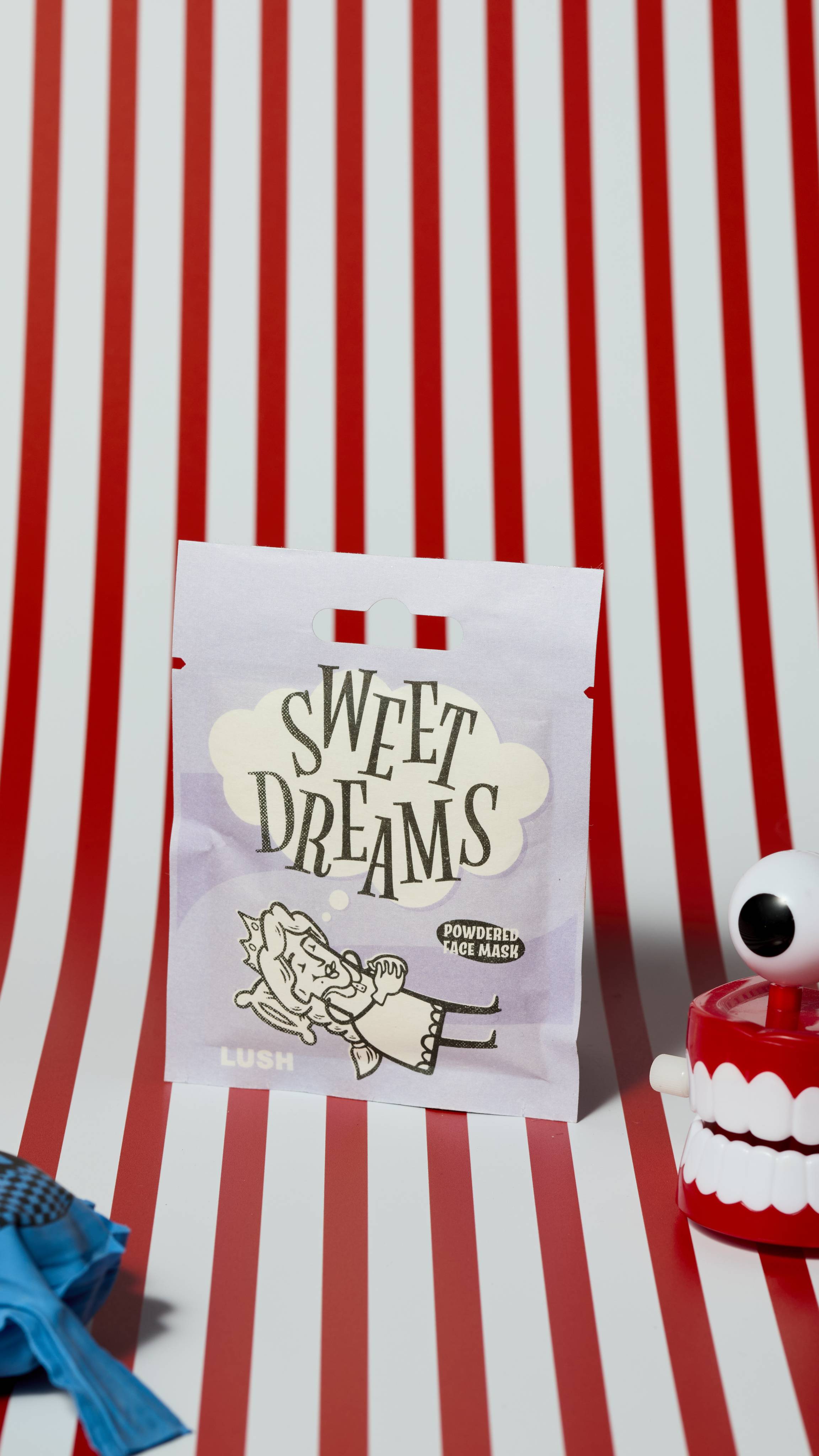 The image shows the Sweet Dreams powdered face mask product sachet on a red and white, vertically striped background. There is a whoopie cushion and wind-up chattering teeth that are just visible.