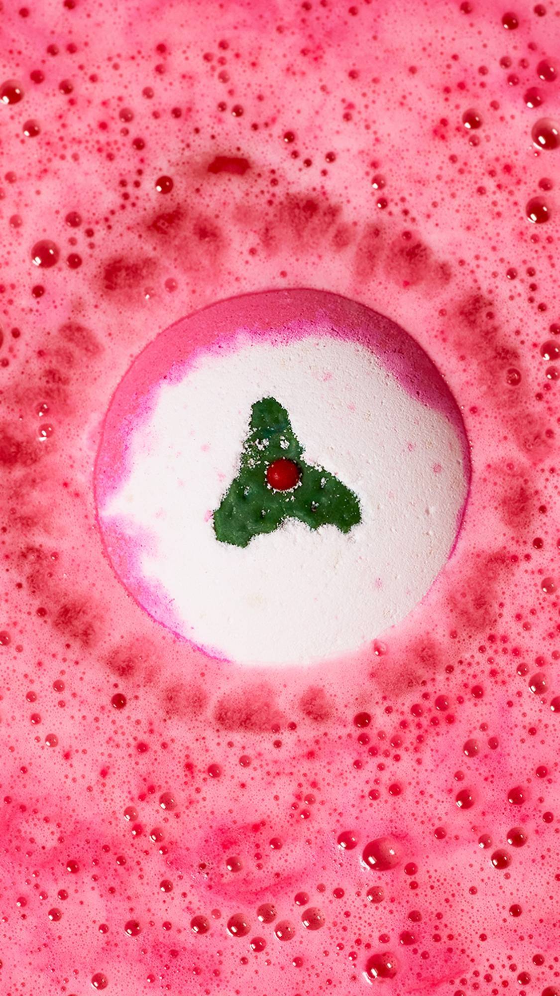 An aerial shot of the perfectly pink Sweet Pudding festive bath bomb. The product is sitting on the water as it gives off a thick velvet blanket of deep pink foam. The decorative holly leaf and berries are visible on top. 