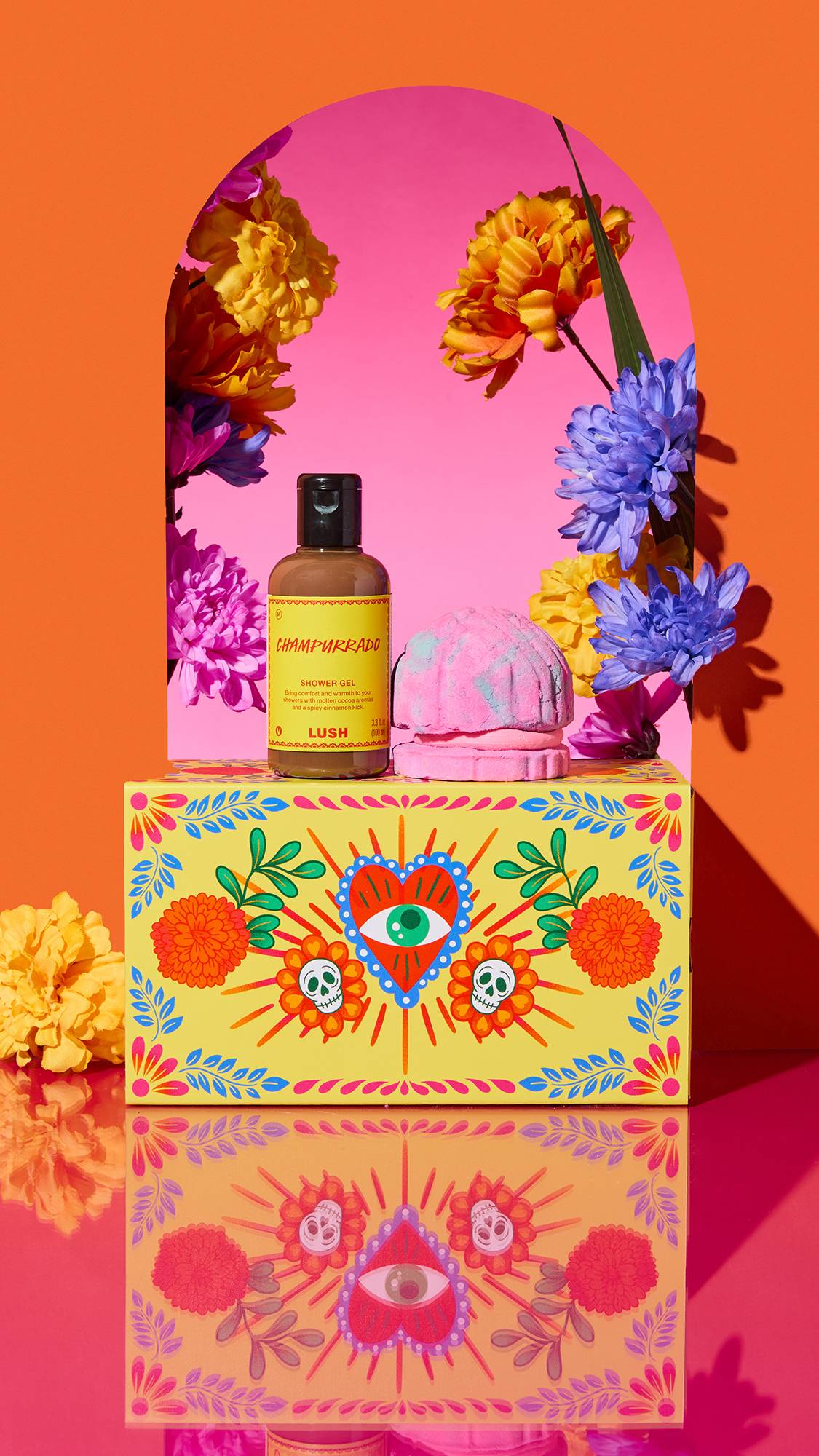 The image shows the 'Sweet Recuerdo' gift set framed by an orange arch and colourful flowers. A folk-art box sits on a reflective pink surface, with a Champurrado shower gel bottle and a Concha Cubbleroon bubble bar on top.