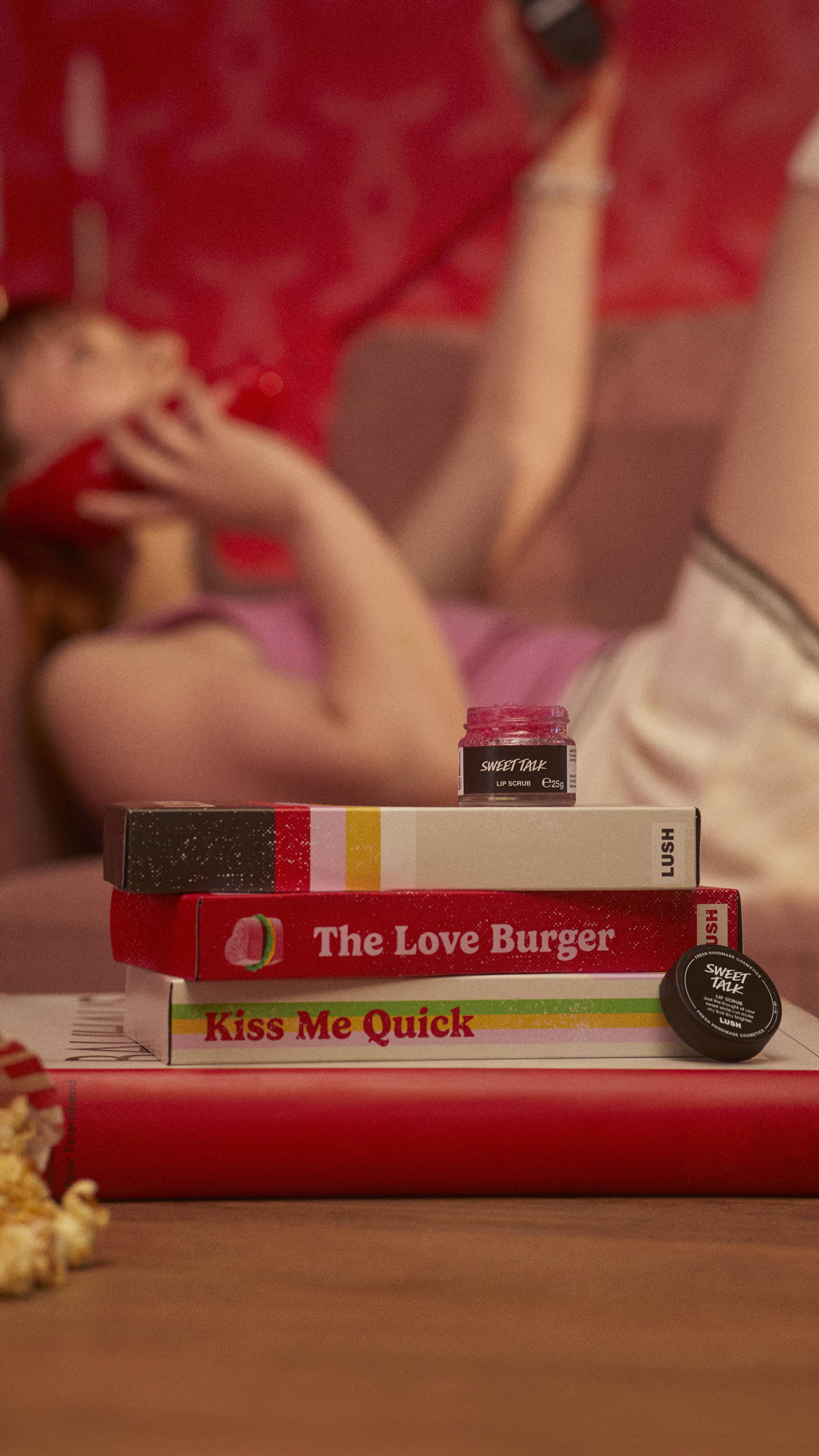 The Sweet Talk lip scrub pot is on a stack of VHS tape boxes as the model is in the background with a retro corded phone.