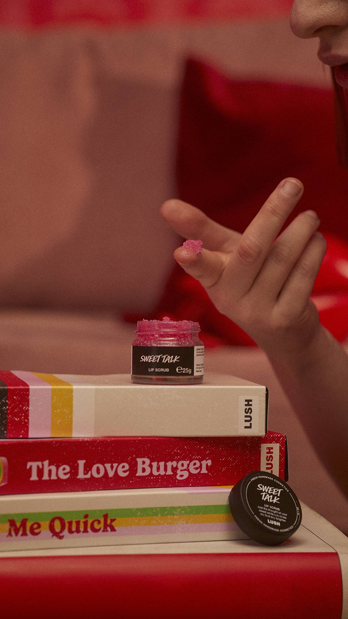 The Sweet Talk lip scrub pot is on a stack of VHS tape boxes as the model scoops out some product onto their fingertip.