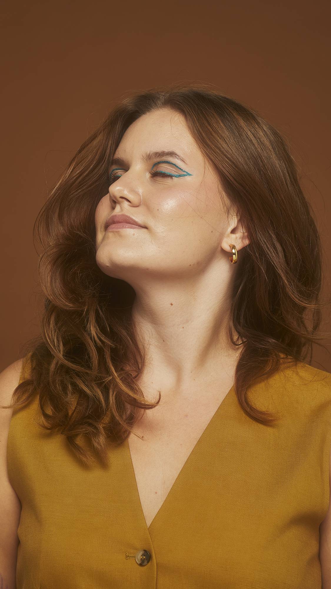 Model wears bold teal eyeliner showing healthy, thick, chestnut hair on an earth-toned background.