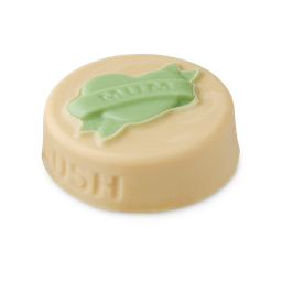 Symbol of Love. A flat, circular, ivory-coloured solid balm with the word LUSH embossed on the side. On top is a pale green heart with a ribbon across which reads "MUM".