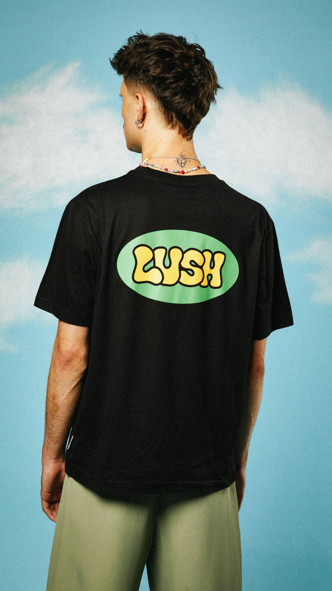 Image shows the back of the t-shirt as the model faces away. The t-shirt back has a large, central retro LUSH logo. 