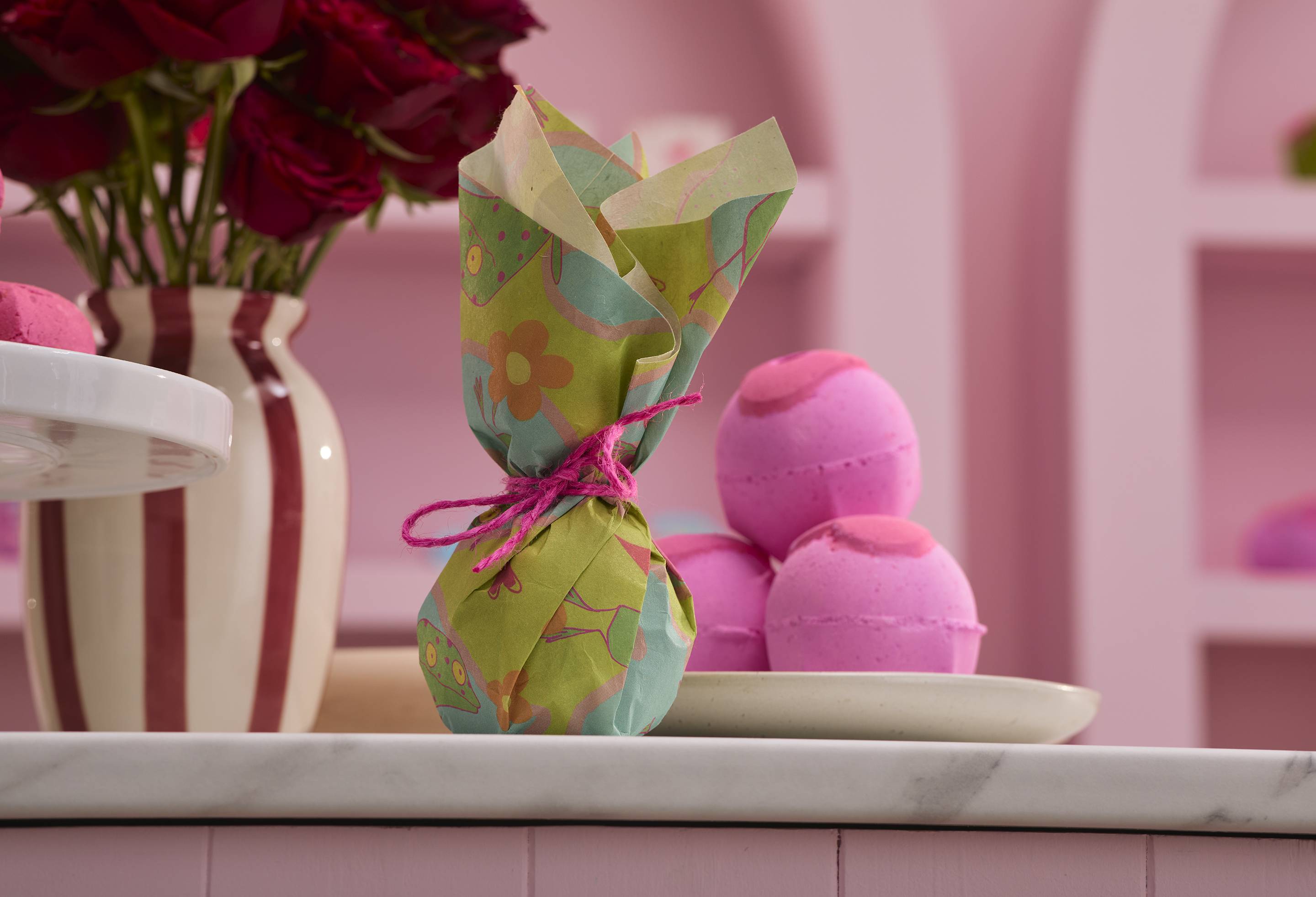 The image shows a pink kitchen background with a striped vase of roses and a plate of piled bath bombs. The Take the Leap lokta paper has been neatly wrapped around a bath bomb-shaped gift and tied with pink string.