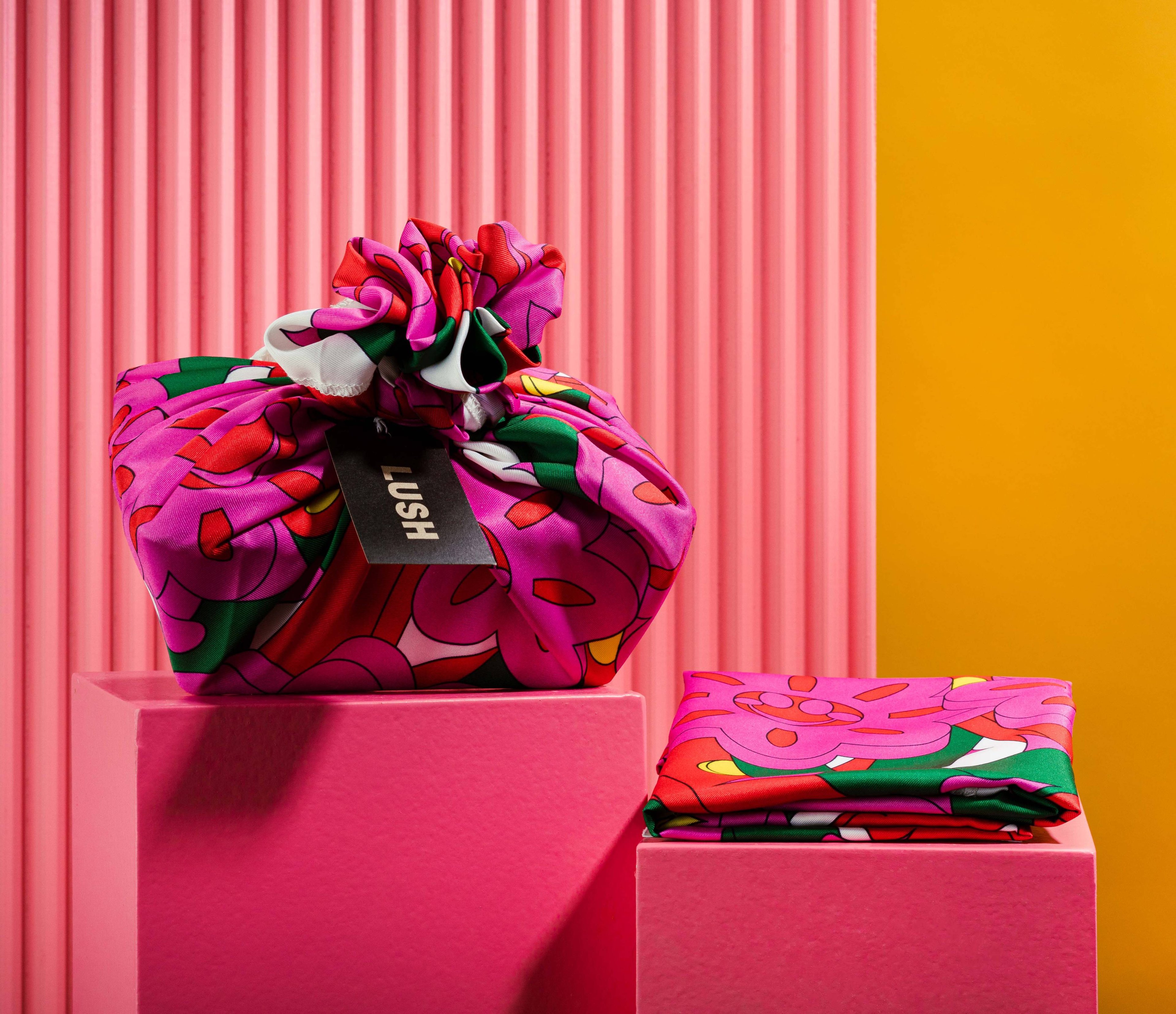 The Knot Wrap is decoratively wrapped with a Lush gift tag, alongside one folded, in front of a pink and yellow background. 