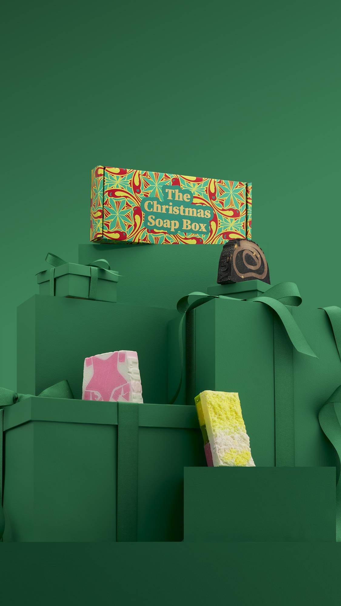 The image shows a large pile of differently-sized wrapped green gifts with ribbons, on a matching green background. The colourful Christmas Soap Box gift box sits on top of the pile, and the three included soaps sit on lower green gifts. 