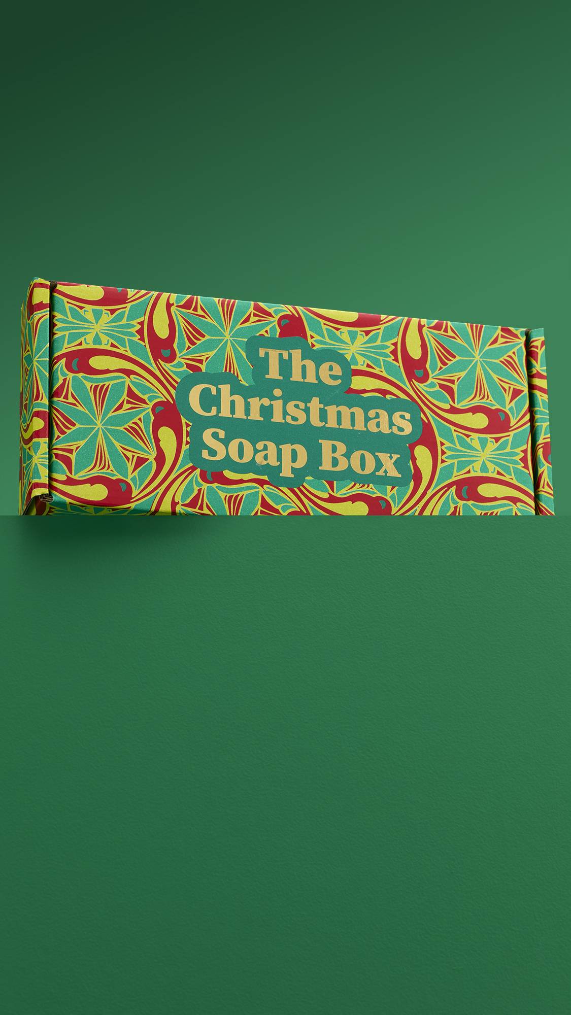 The image shows the vibrant, "The Christmas Soap Box" gift box, sitting on a green platform against a matching green background. 