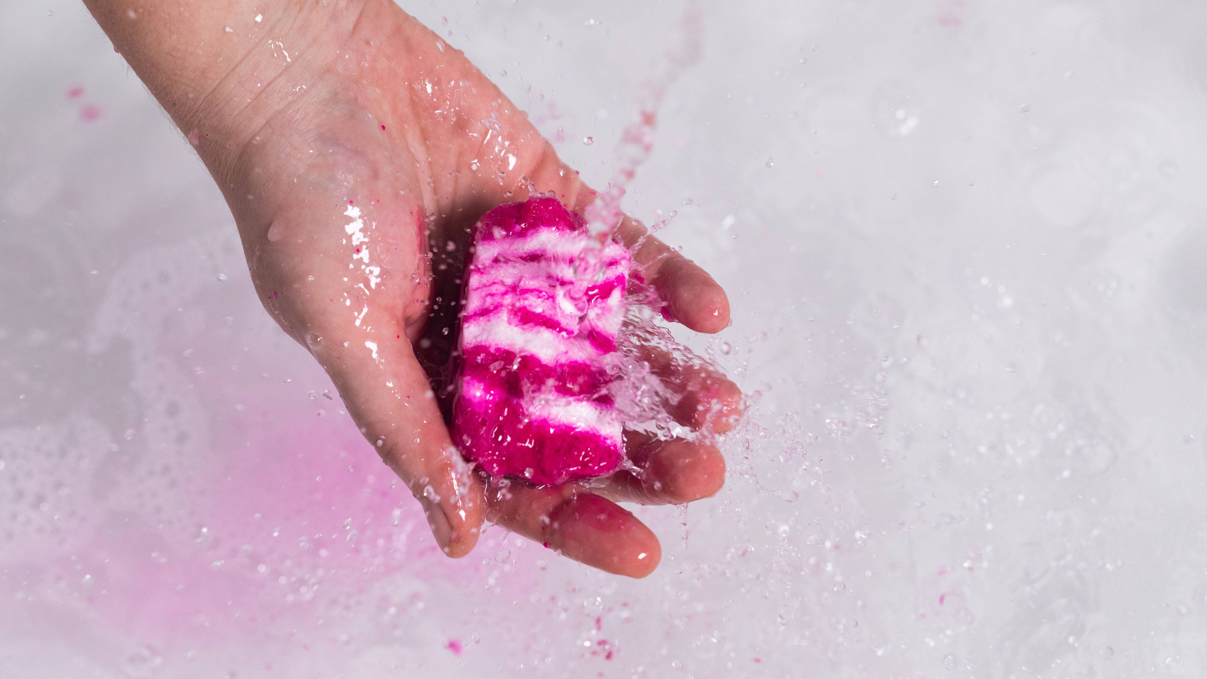 The model holds the Comforter bubble bar under running water, causing pooling, pink splashes of bubbles below.