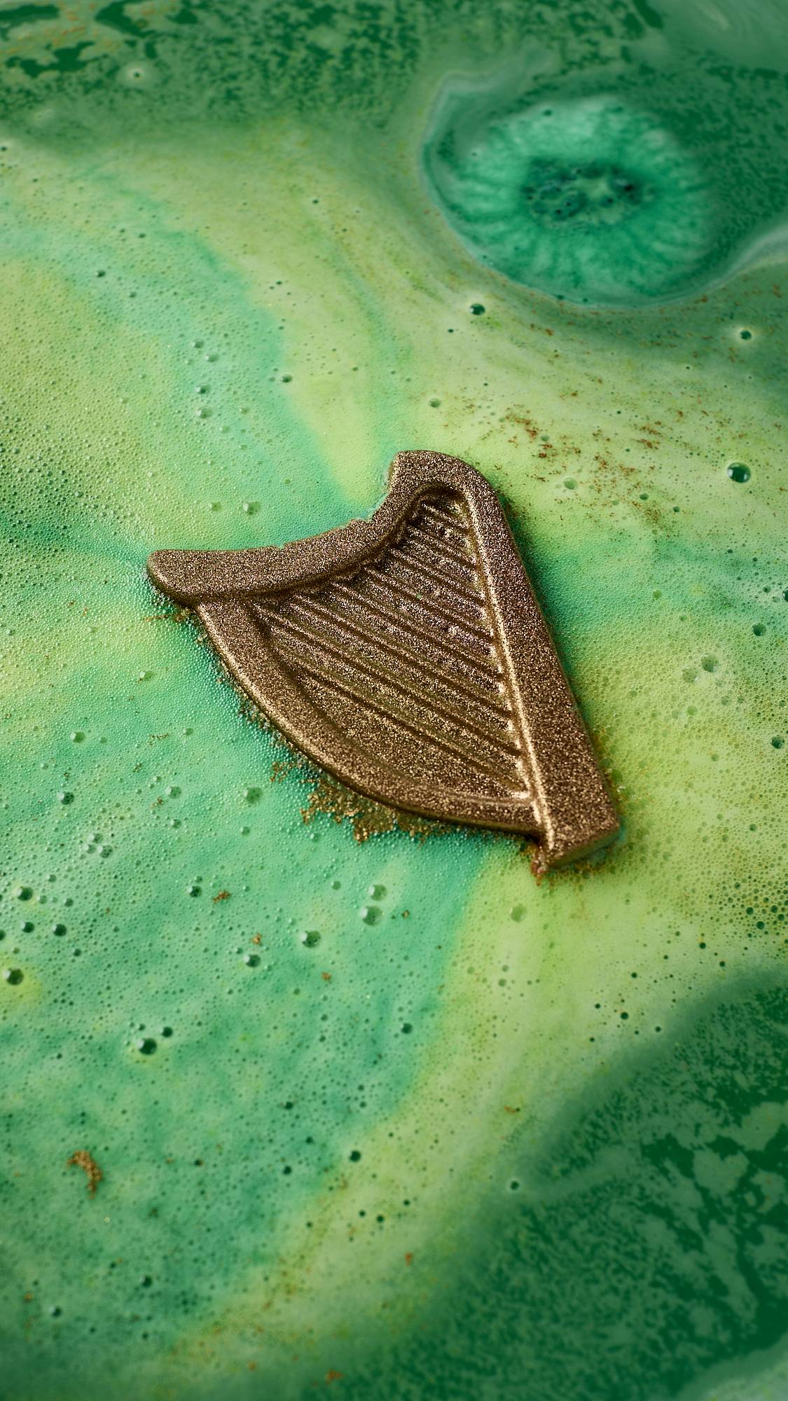 An aerial view of the St. Patrick's Day-themed product, the harp-shaped Craic bath bomb. The product is floating in the centre surrounded by a blanketing layer of thick, velvety, deep green and yellow foam. 