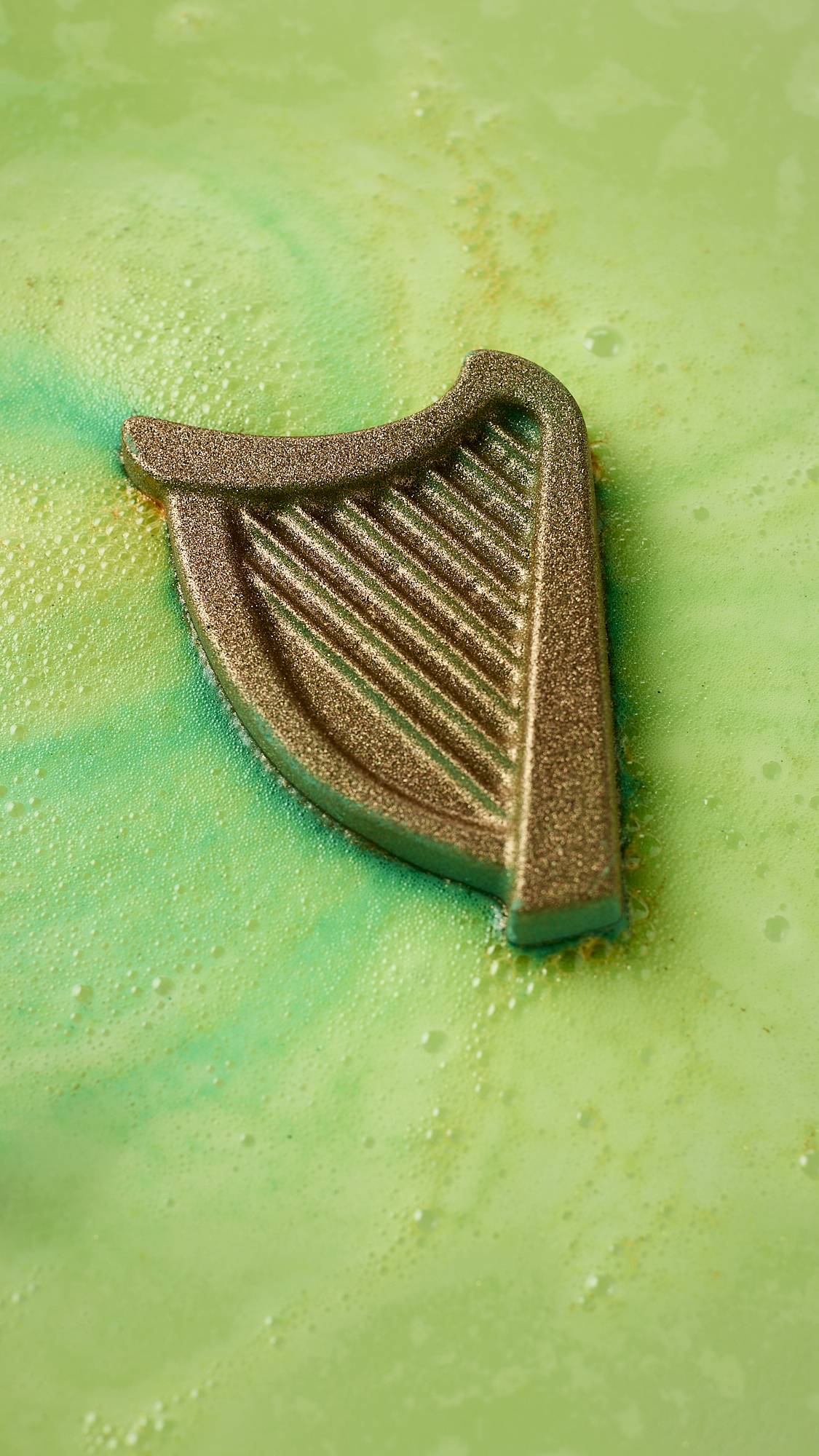 A close-up image of the St. Patrick's Day-themed product, the harp-shaped Craic bath bomb. The product is floating in the centre surrounded by a blanketing layer of thick, velvety, deep green and yellow foam. 
