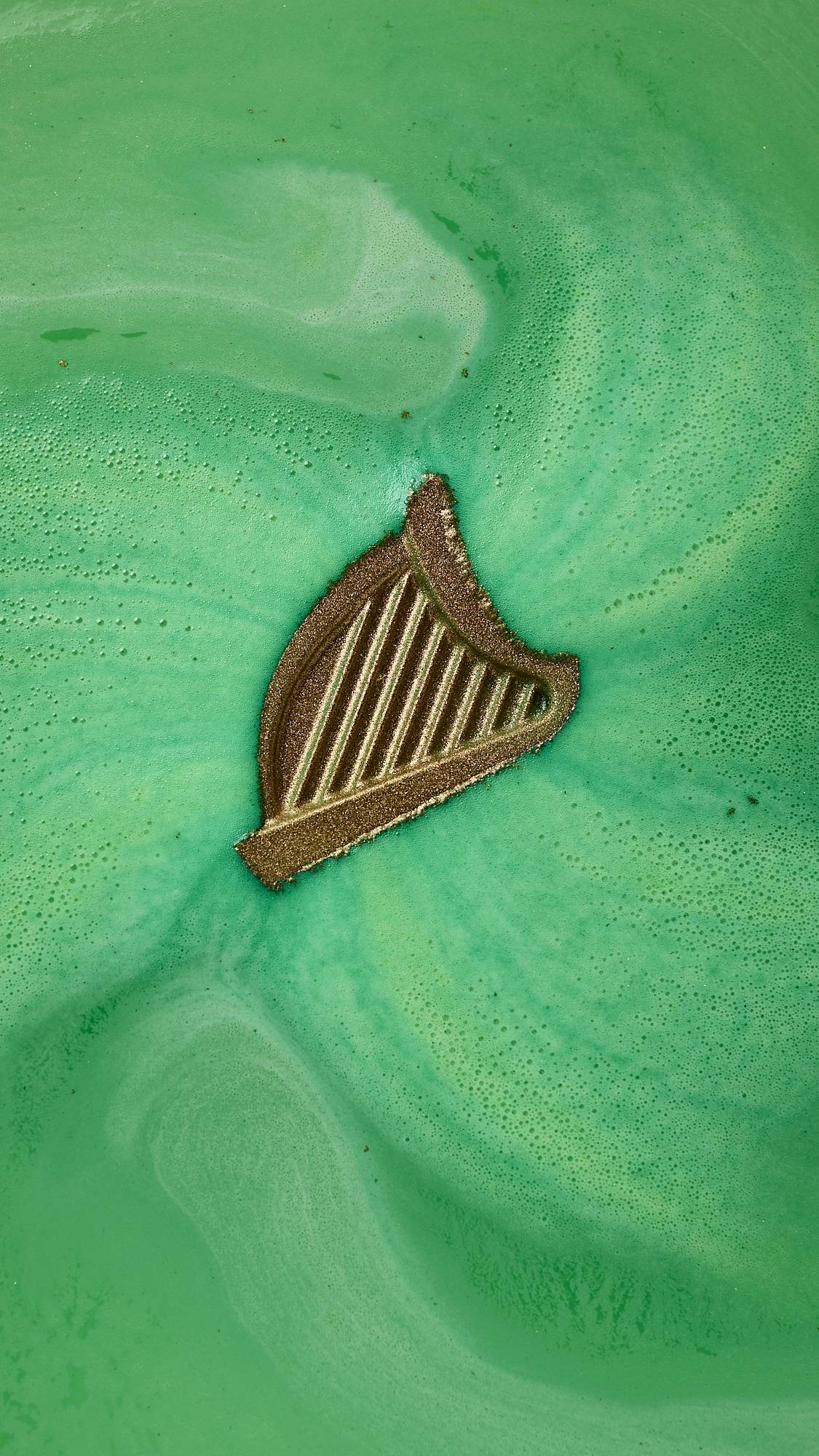 An aerial view of the St. Patrick's Day-themed product, the harp-shaped Craic bath bomb. The product is floating in the centre surrounded by a blanketing layer of thick, velvety, deep green foam. 