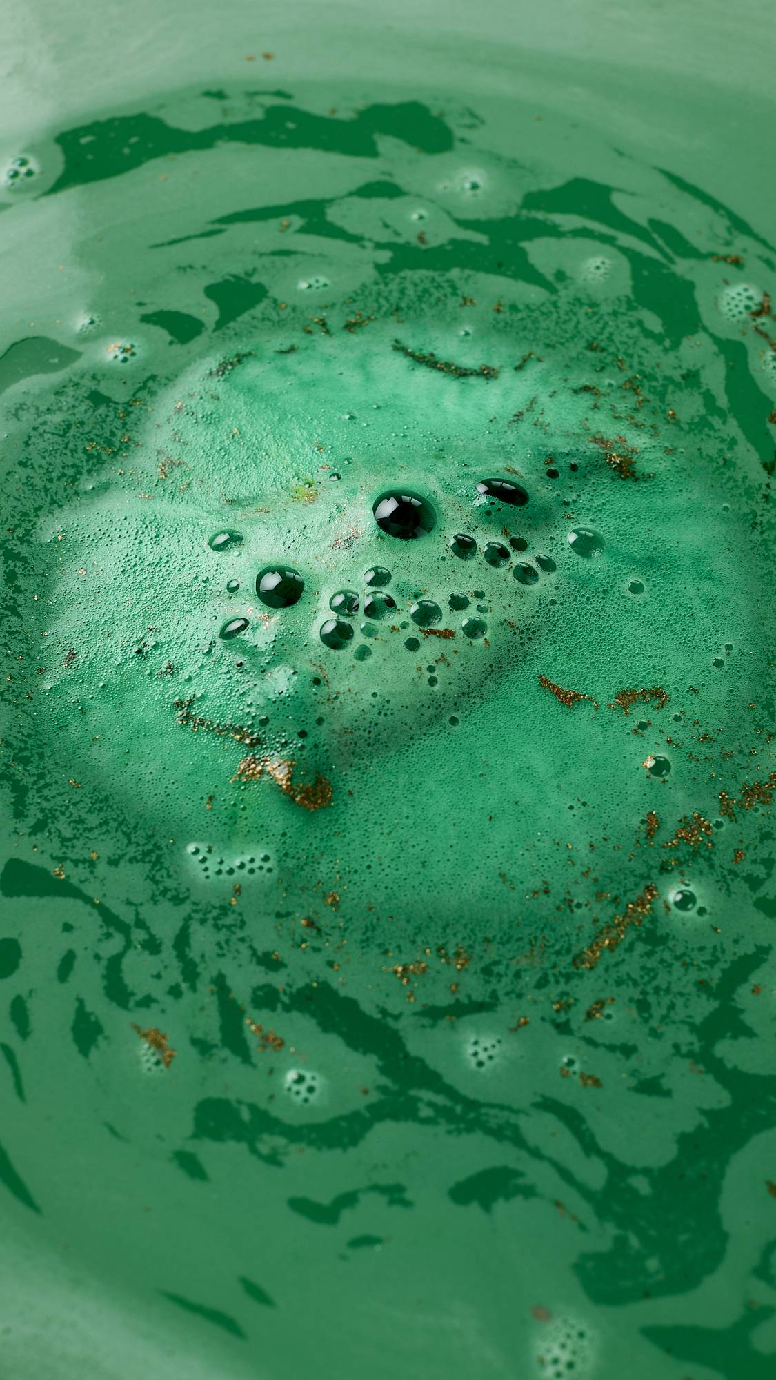 An aerial image of the bath water after using the St. Patrick's day-themed, harp-shaped Craic bath bomb. The bath has been turned a deep green colour with a layer of thick, velvety foam laced with flecks of golden glitter. 