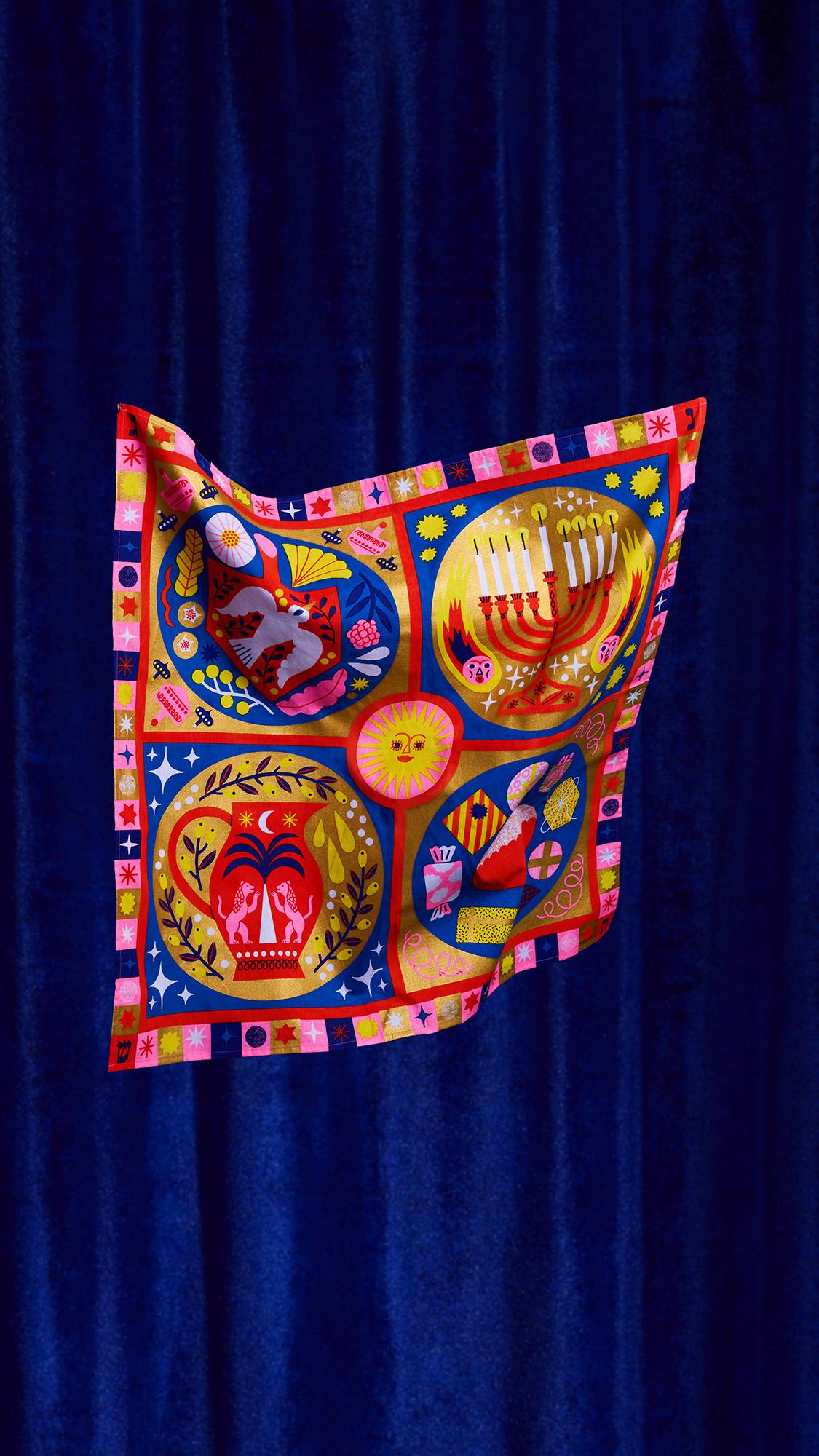 The Eighth Flame Knot Wrap floats diagonally against royal blue velvet. The colorful fabric features four main festive motifs: a menorah, a dove, an oil pitcher, and various gifts and sweets.