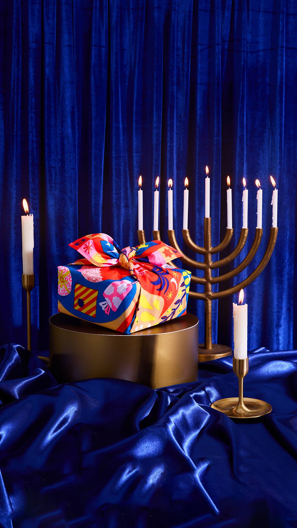 A colorful gift, wrapped in the The Eighth Flame Knot Wrap with a bow, sits on a gold stand. A gold menorah with eight lit candles and two single lit candles are visible against a royal blue velvet and satin backdrop.