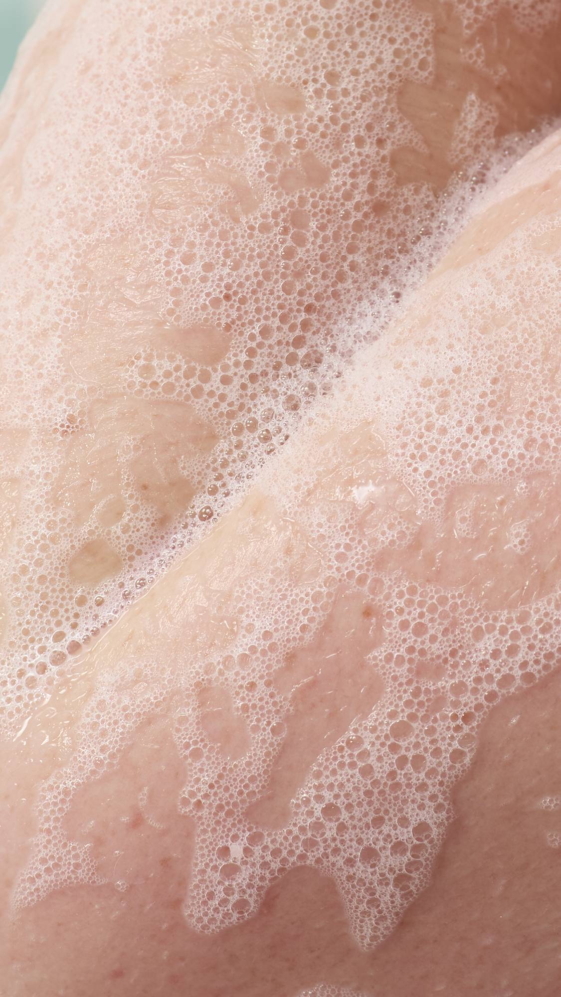 A close-up of a person's skin covered in a light, airy lather of white soap bubbles. The delicate foam spreads across the skin's surface, reflecting a soft, clean texture during a bath.
