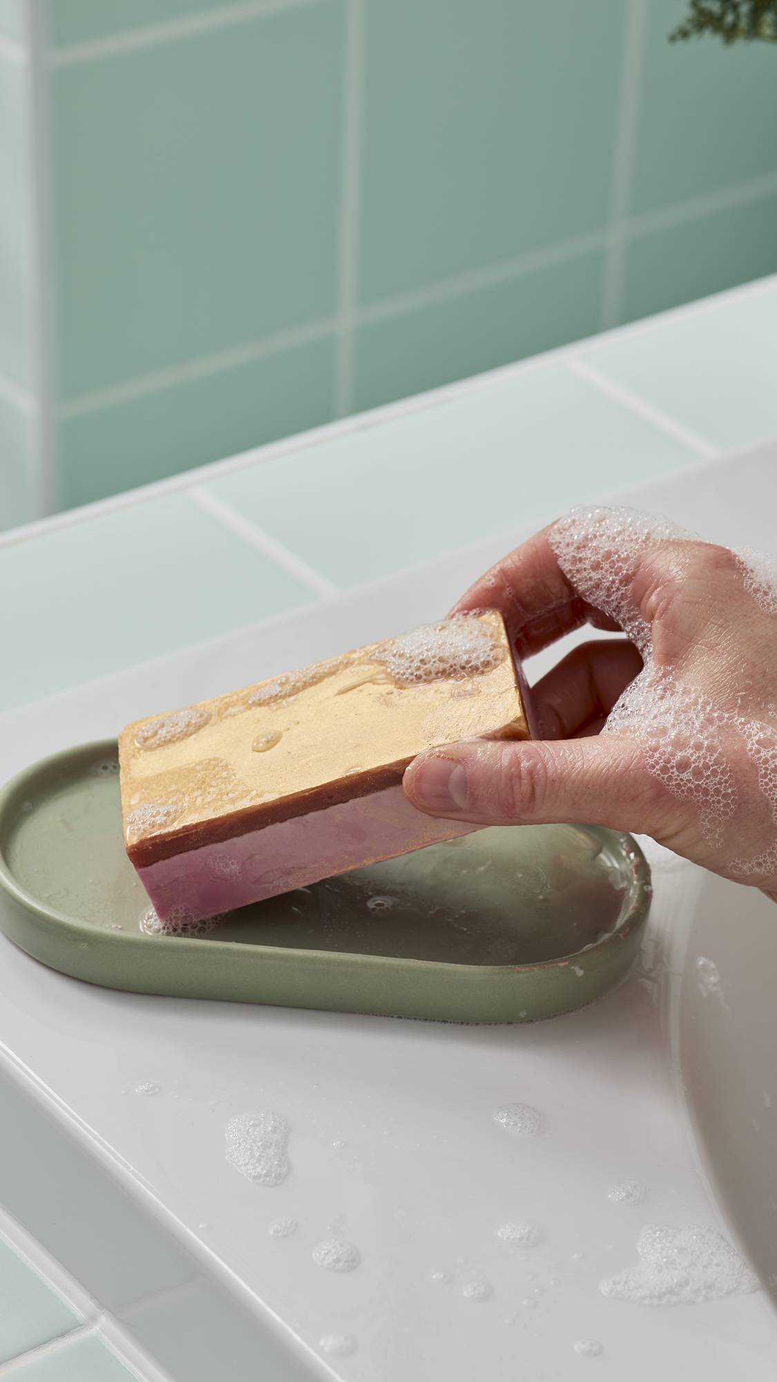 A hand picks up a rectangular soap bar with a shimmering gold top and pink base from a green soap dish on a white tub ledge. Small white bubbles cling to the gold surface and the person's hand.