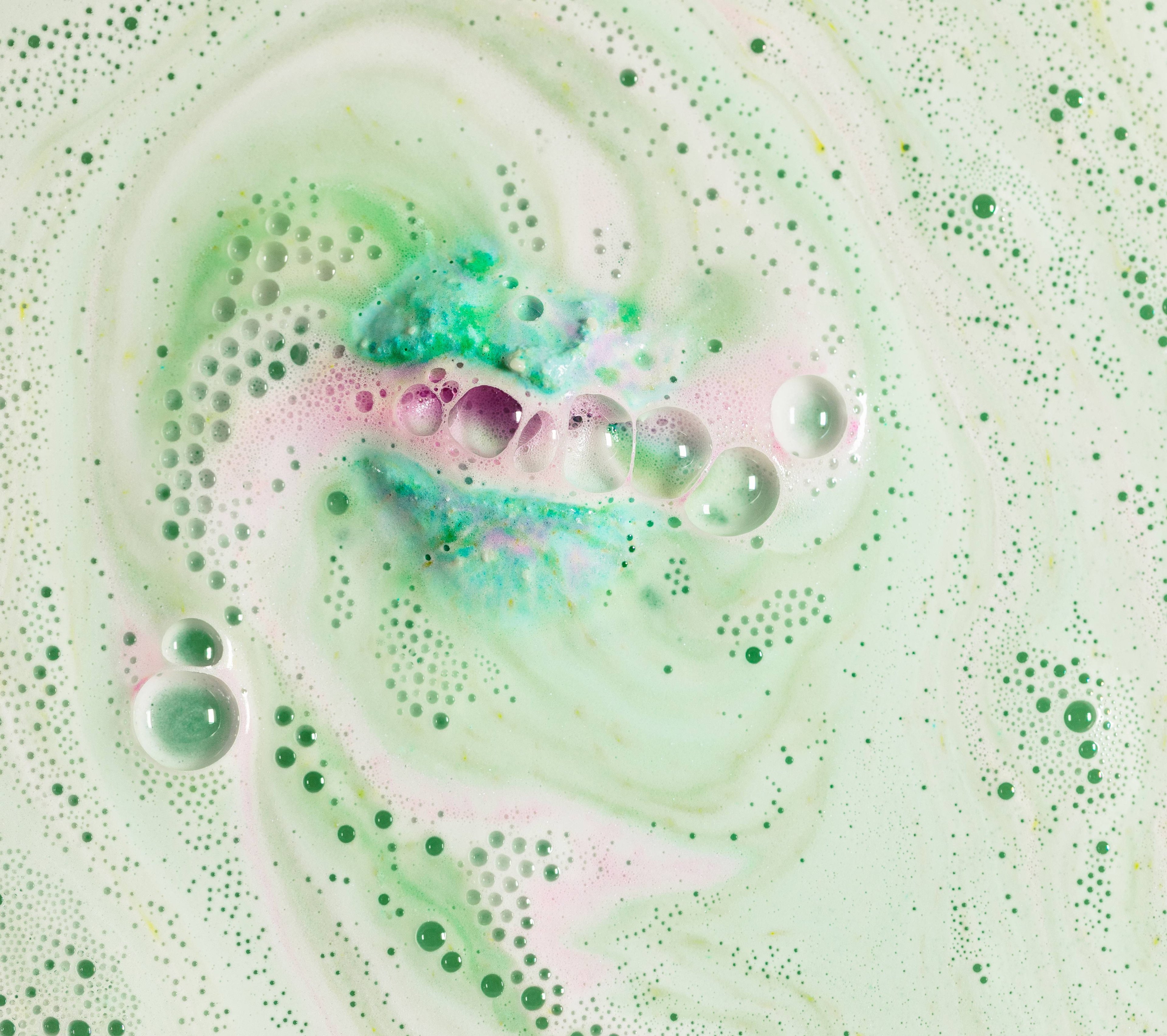 A spiral of pastel pink and green foam and bubbles emerge in a chain from a crack in the floating Lakes bath bomb.