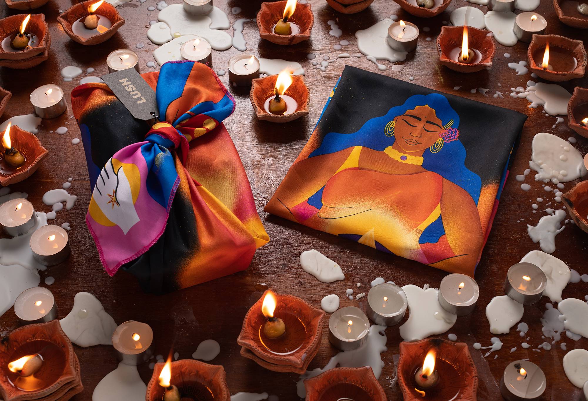 The knot wrap is shown folded and tied around a square package, laid on a wooden surface surrounded by lit tealight candles.