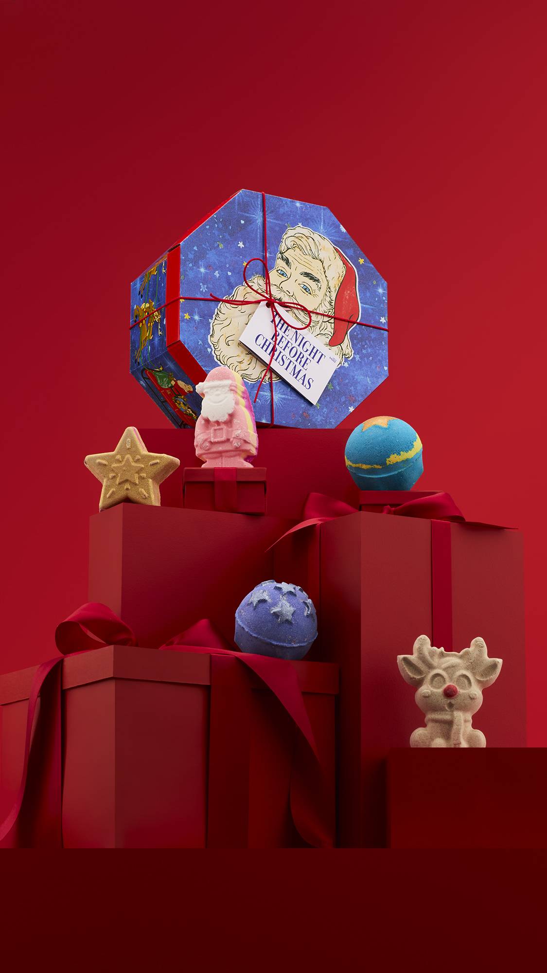 The image shows a large pile of differently-sized wrapped red gifts with ribbons, on a matching red background. The octagonal Night Before Christmas gift box sits on top of the pile, and the five included bath bombs sit on lower red gifts. 
