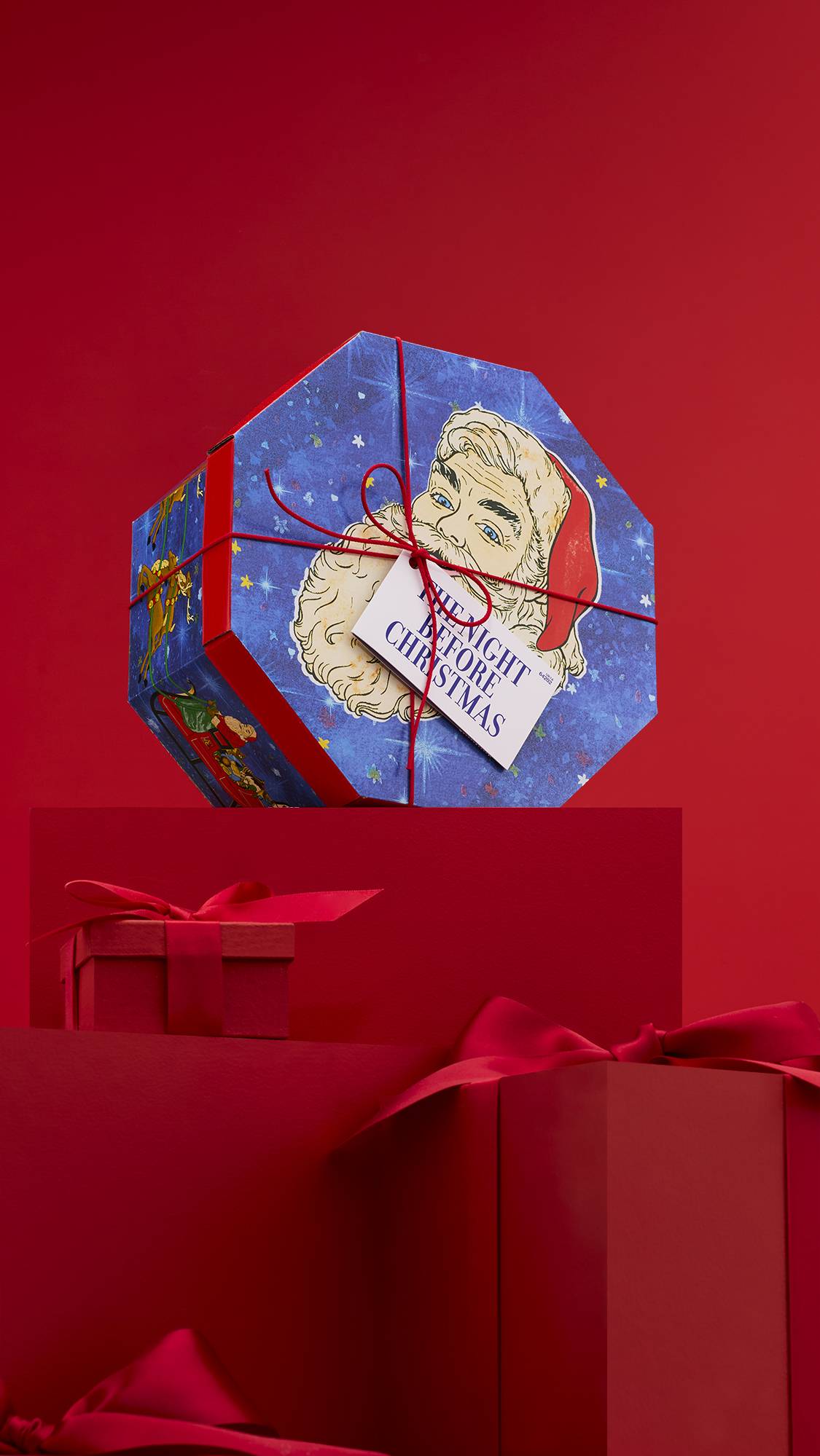 The image shows the octagonal Night Before Christmas gift box, tied with a red string and a white gift tag, sitting on a solid red platform against a matching red background. 