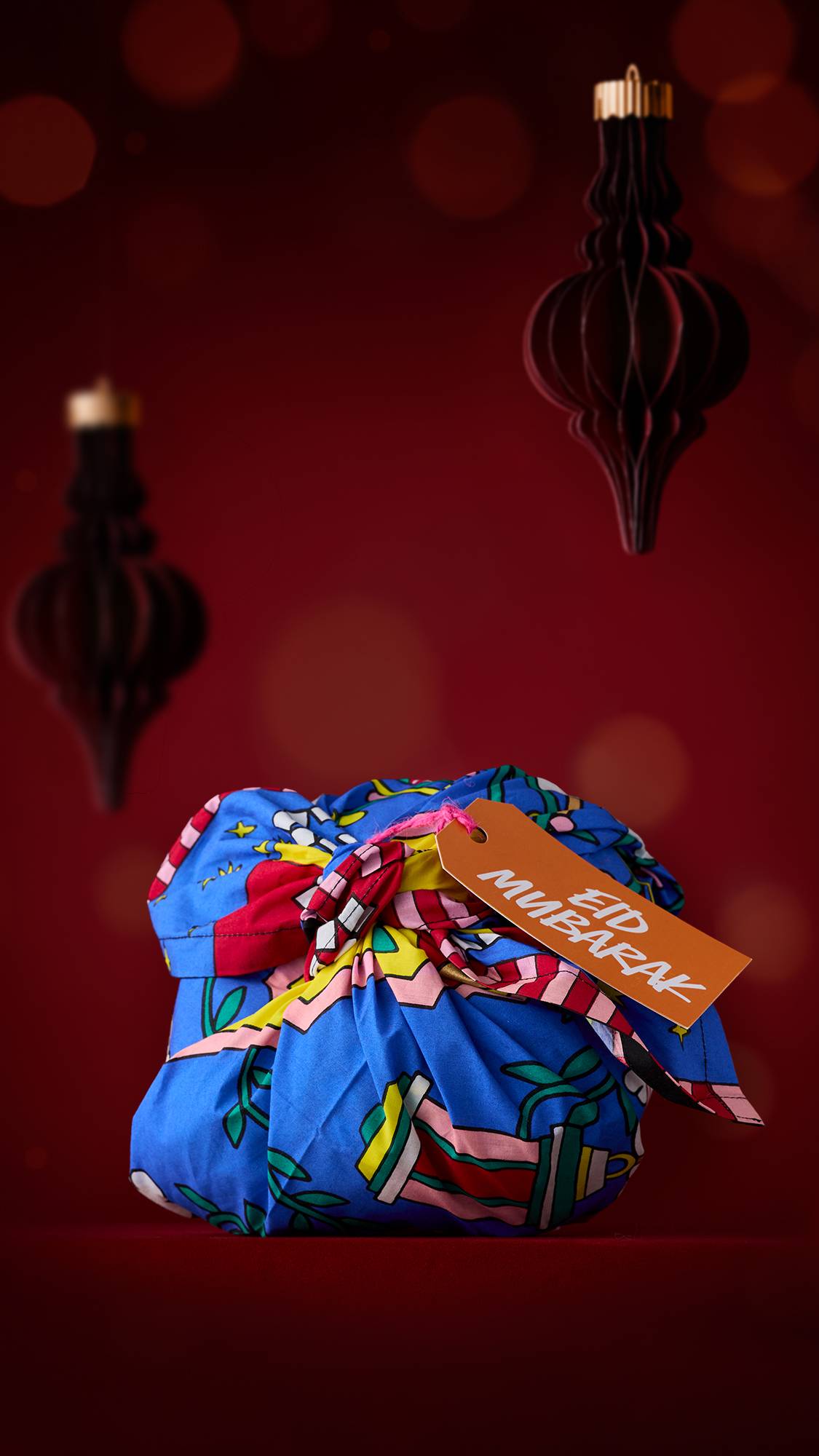 The image shows a gift wrapped in a blue floral "The Night Before Eid" Knot Wrap with an orange "Eid Mubarak" tag. The gift sits on a dark red surface with two lantern-shaped ornaments hanging in the blurred background.