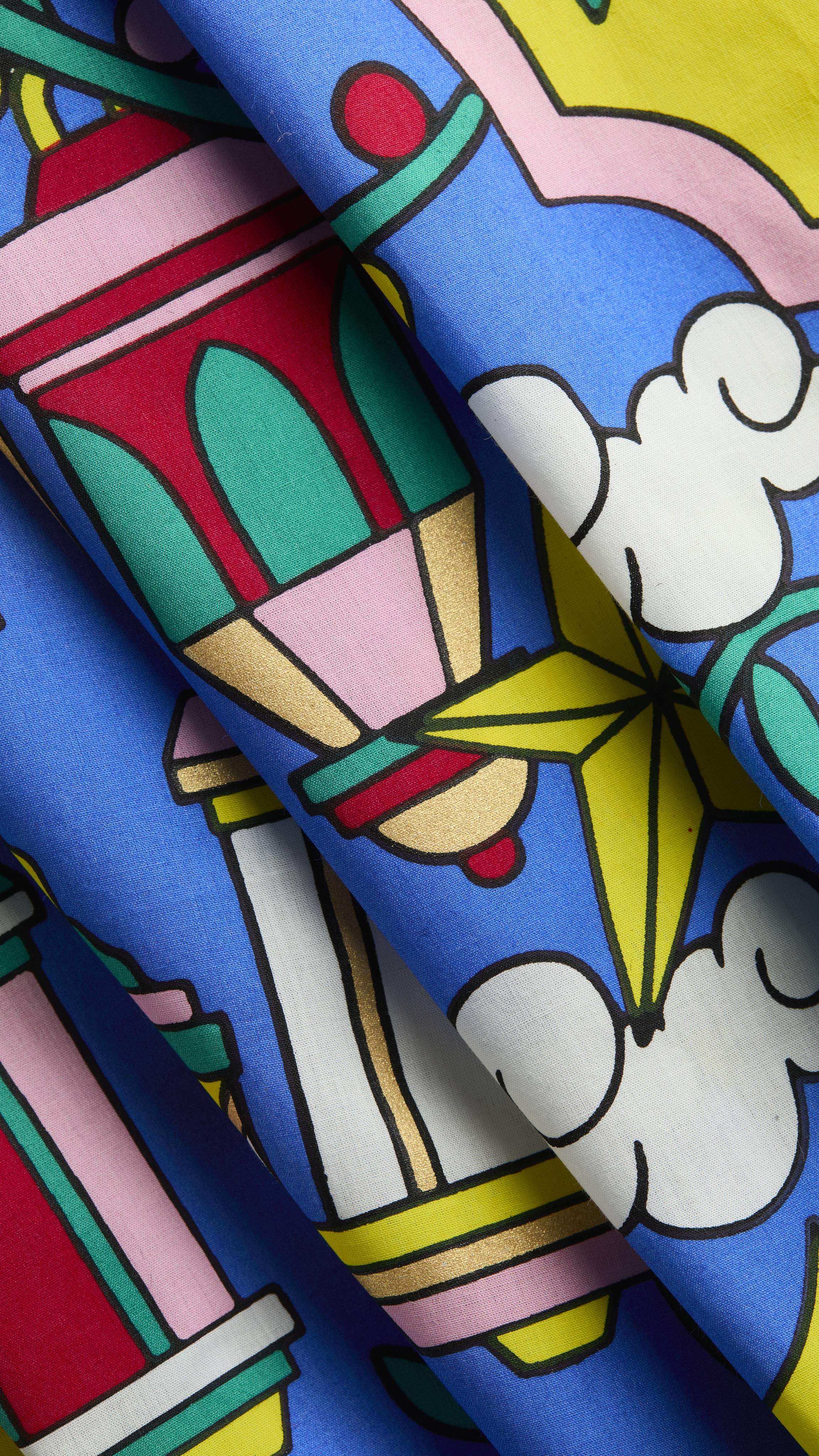 A close-up of a blue fabric "The Night Before Eid" Knot Wrap featuring a vibrant illustration of a colourful lantern, yellow stars, and white clouds. The material is folded diagonally, highlighting the crisp print and smooth texture.