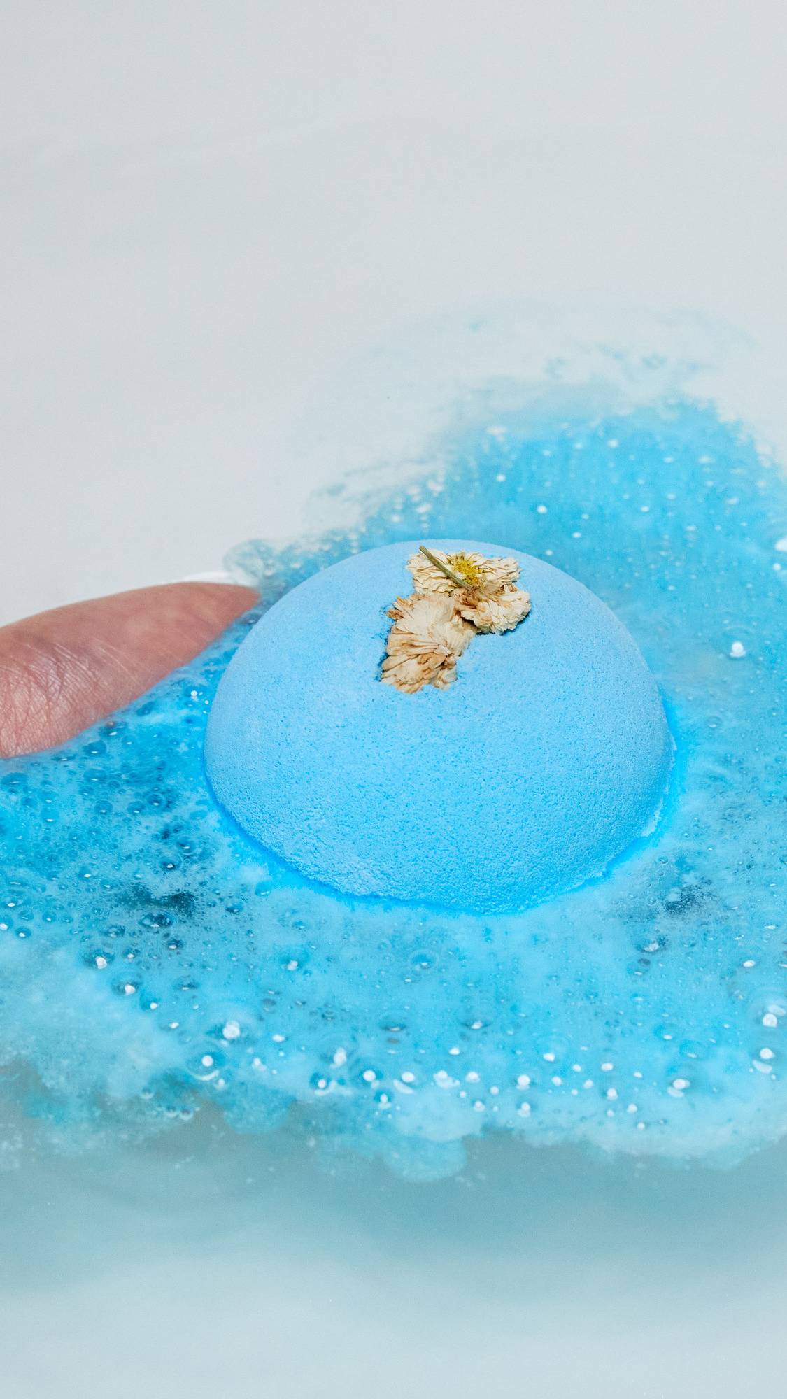 The One With Chamomile bath bomb is being placed into the bath and is slowly dissolving creating a sea of deep blue water and thick, velvety blue foam. 