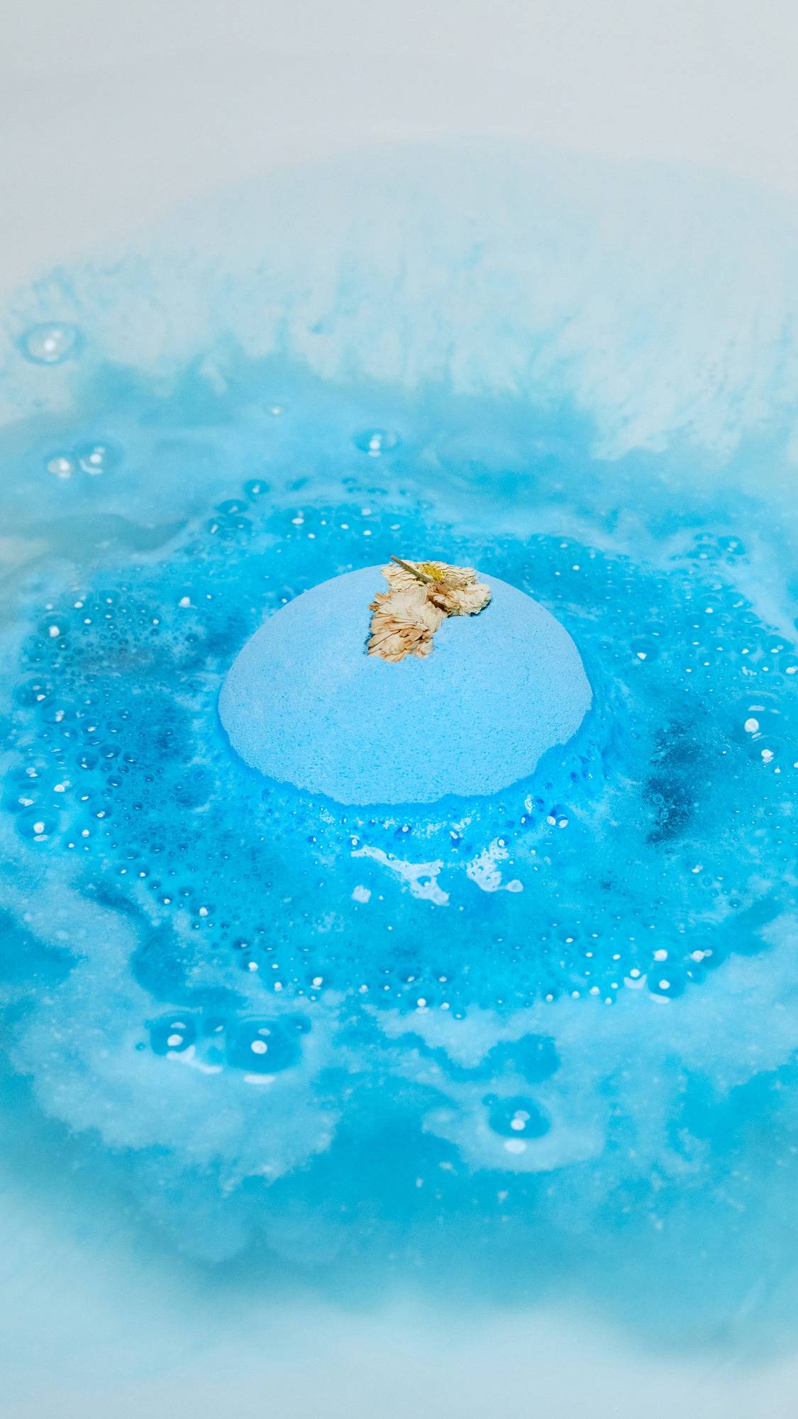 The One With Chamomile bath bomb is slowly dissolving creating a sea of deep blue water and thick, velvety blue foam. 