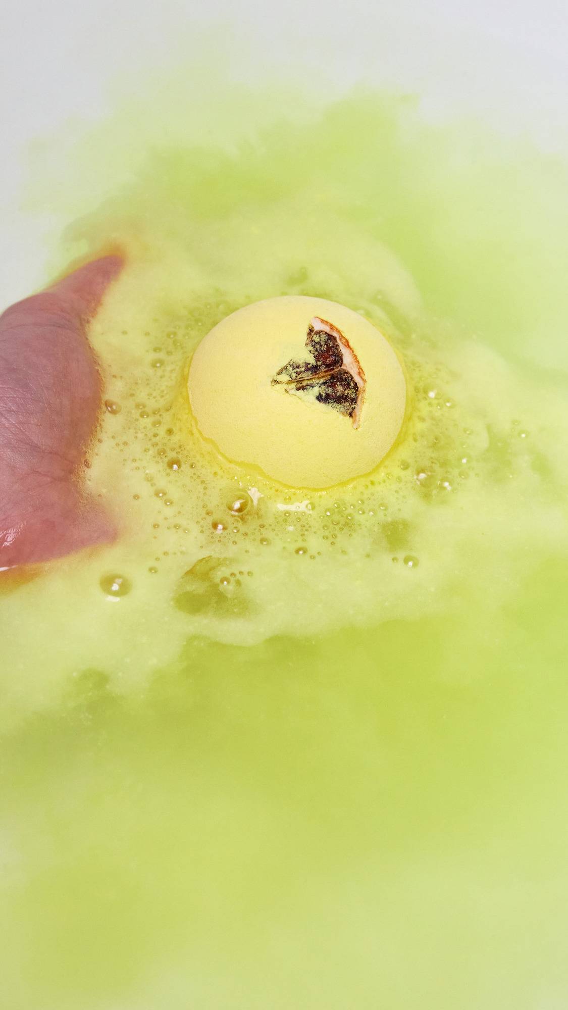 The One With Lemon Oil bath bomb is being gently placed into the bath by the model and is already giving off delicate, velvety, yellow foam across the surface. 