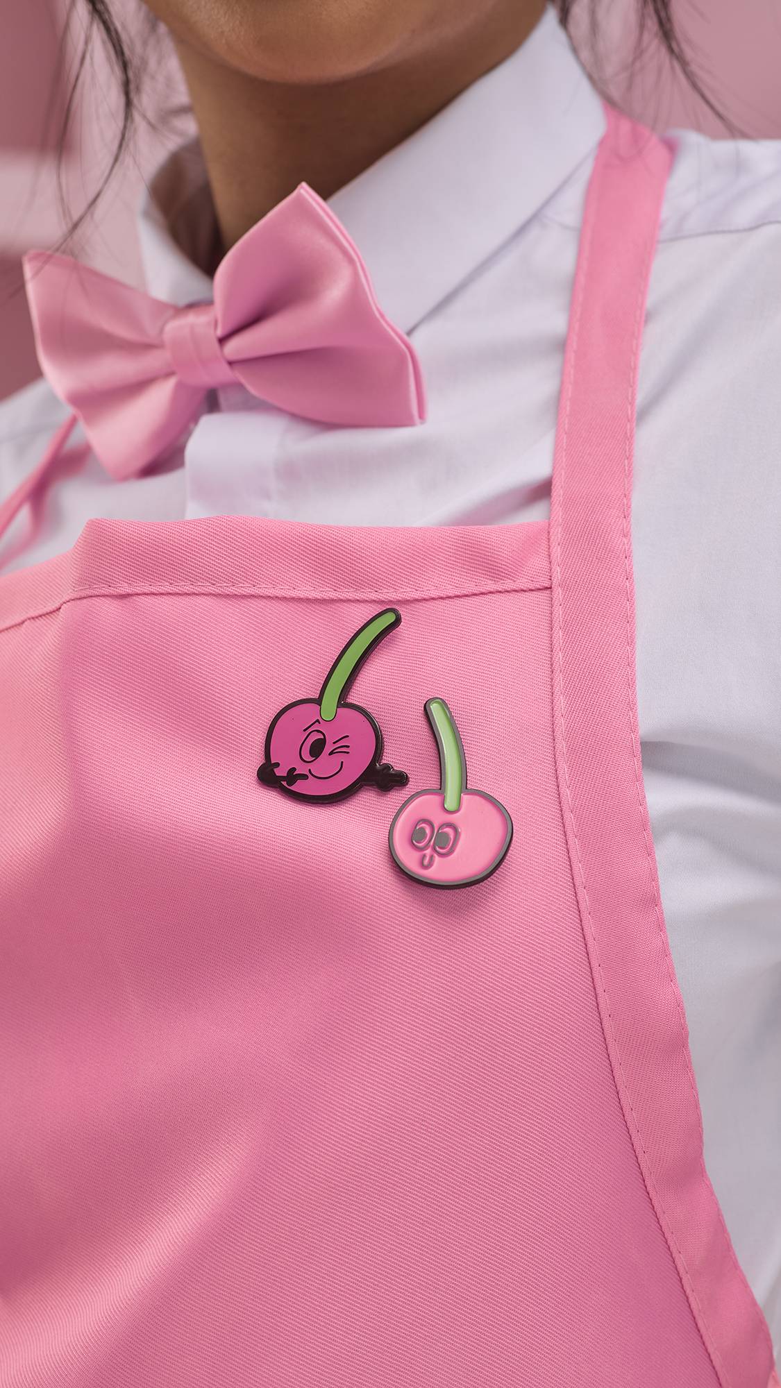 The image shows a close-up of the model as they are wearing a white shirt with a pink bow tie and apron. Fastened to the top of the apron are two halves of the smiling cherry pin badges from the Perfect Paid gift card.