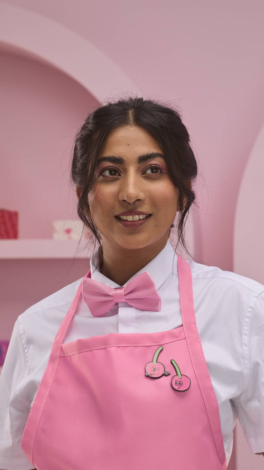 The image shows the model as they are wearing a white shirt with a pink bow tie and apron. Fastened to the top of the apron are two halves of the smiling cherry pin badges from the Perfect Paid gift card.