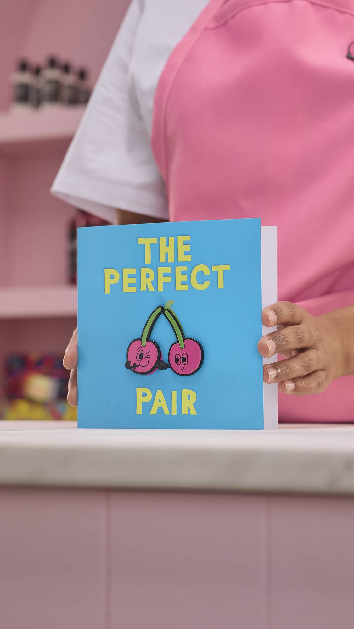 The image shows the model wearing a pink apron and placing The Perfect Pair gift card with detachable cherry pin badges onto a counter. 