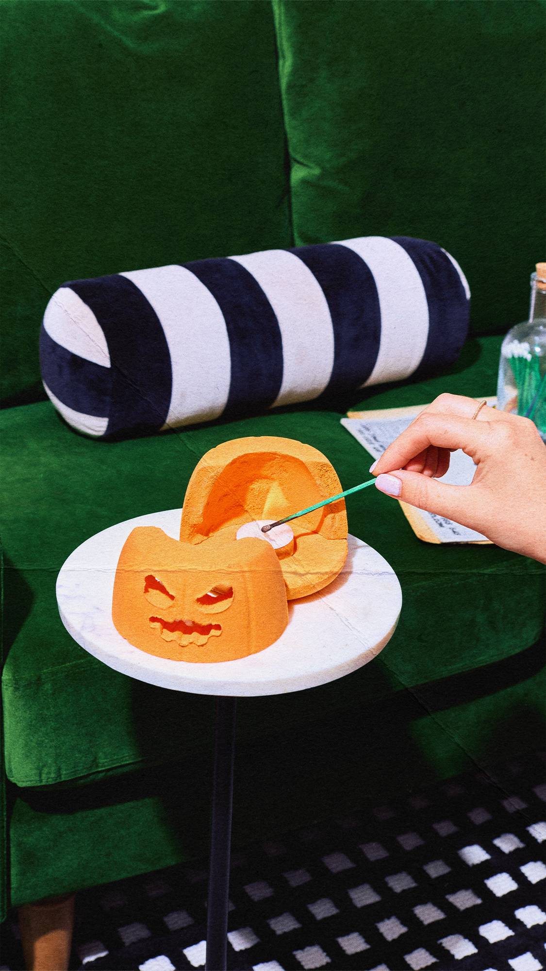 A hand uses a long green match to light a tea light candle inside an open, orange pumpkin-shaped bath bomb diffuser on a small round white table. A green velvet sofa and black-and-white striped bolster cushion sit in the background.