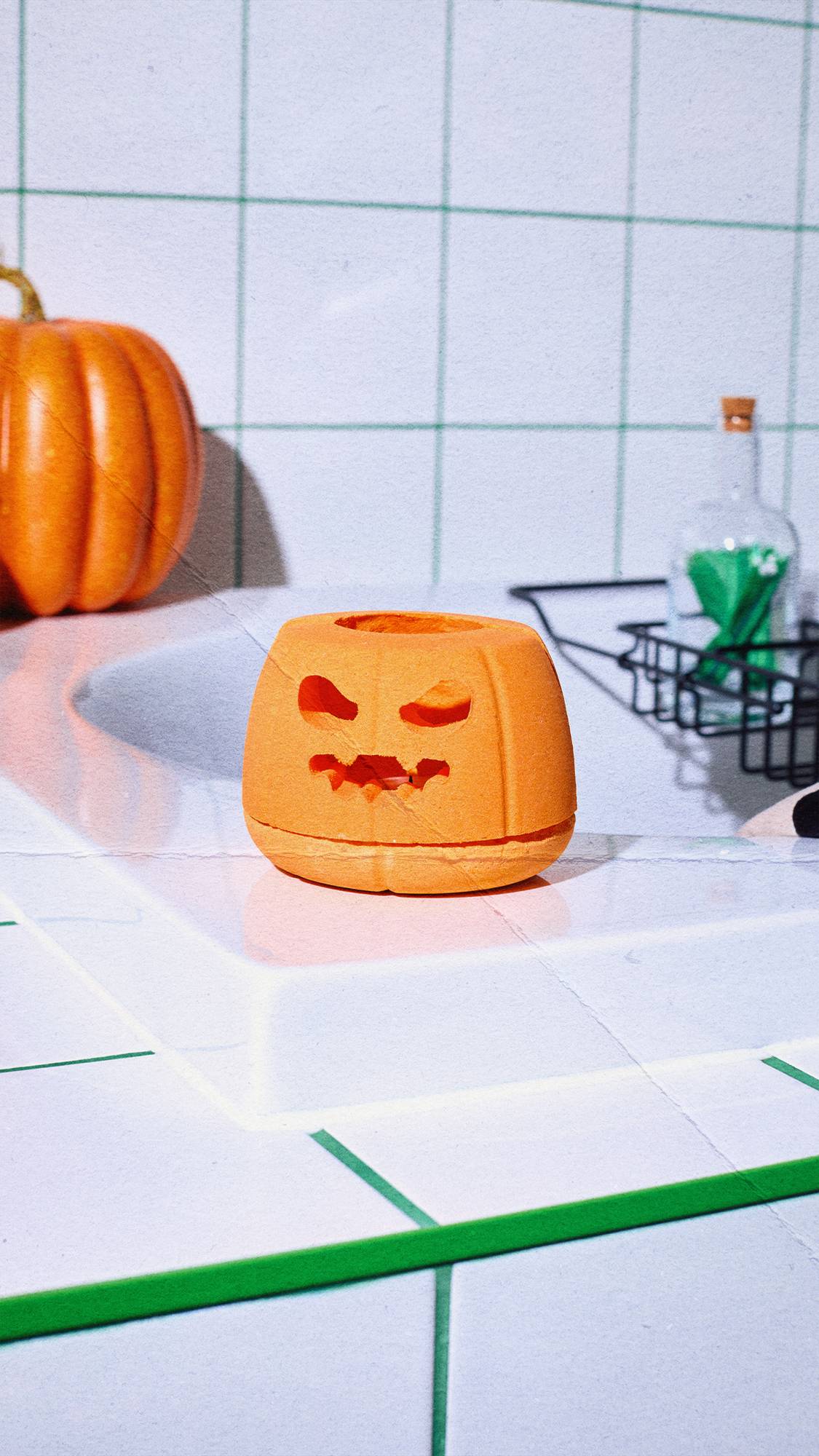 An orange pumpkin-shaped bath bomb diffuser sits on the edge of a white tiled bath. In the background, a large real pumpkin, white wall tiles with green grout, and a glass bottle filled with green matches are visible.