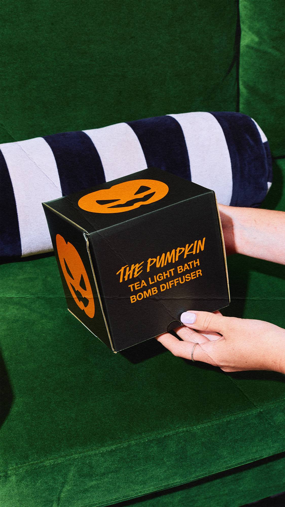 A pair of hands holds a black cube box with orange text reading 'THE PUMPKIN TEA LIGHT BATH BOMB DIFFUSER'. The box features orange jack-o'-lantern faces and is set against a green velvet sofa and striped bolster cushion.