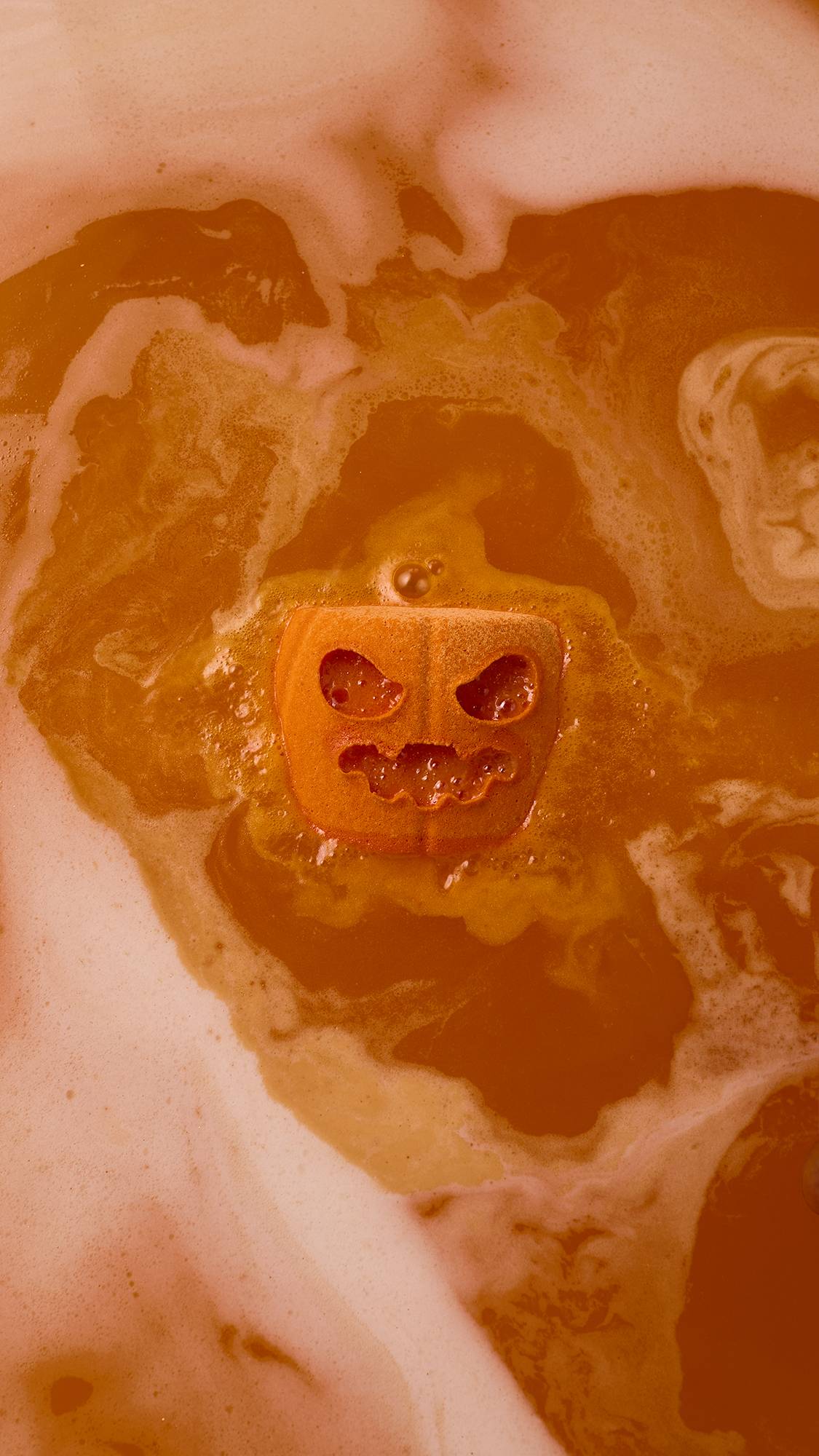 An orange pumpkin-shaped bath bomb dissolves in bathwater, creating vibrant orange water surrounded by swirling white foam.  
