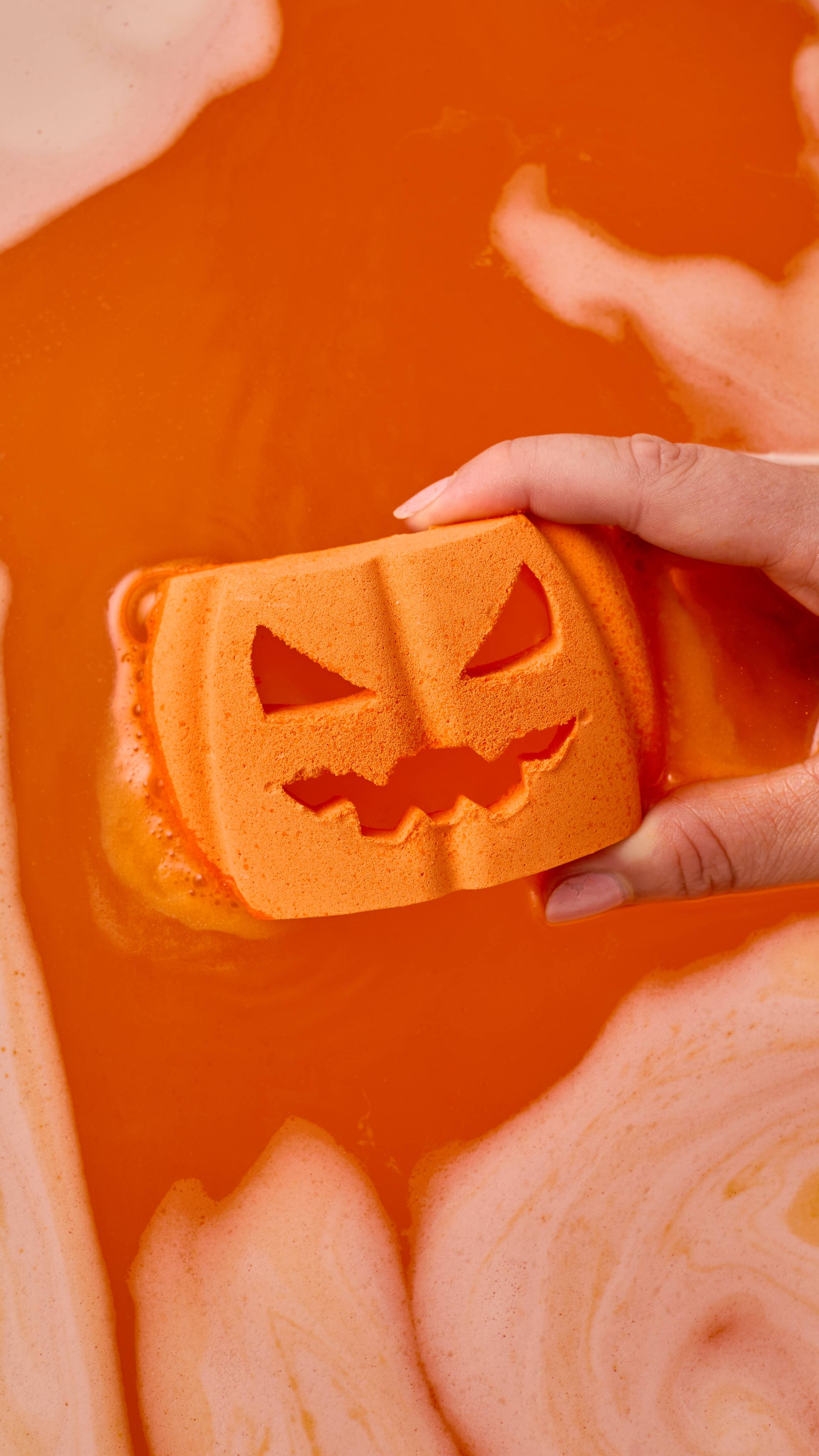 A hand dips an orange pumpkin-shaped bath bomb with a carved jack-o'-lantern face into vibrant orange water. Swirls of thick foam surround the fizzing water.