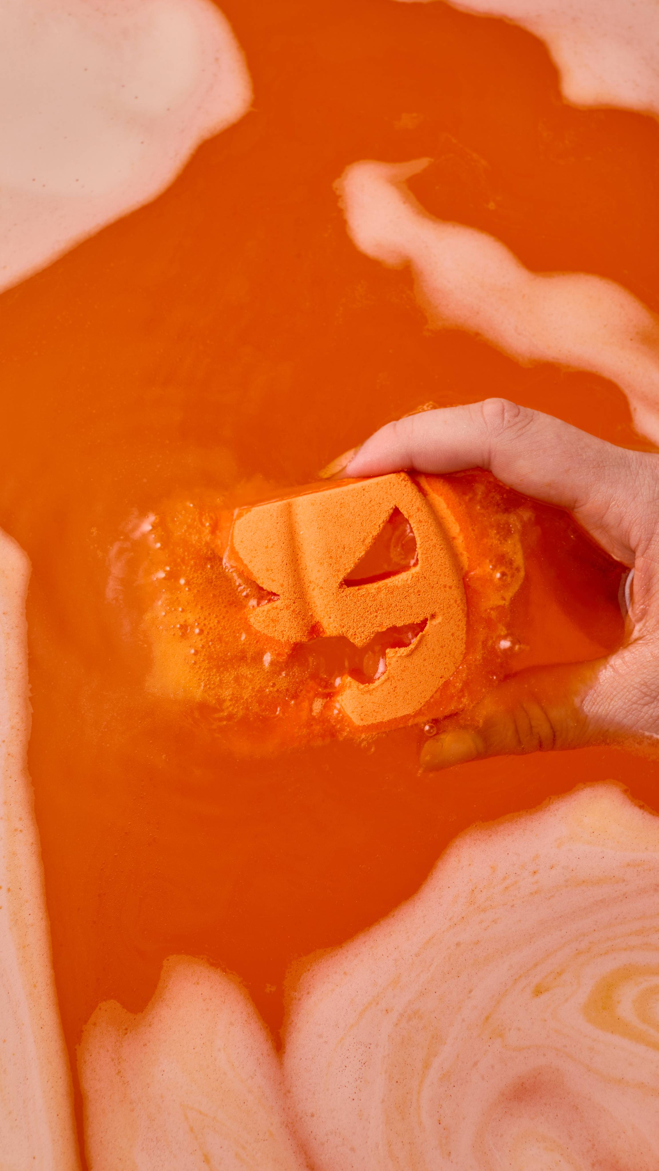 A hand dips an orange pumpkin-shaped bath bomb with a carved jack-o'-lantern face into vibrant orange water. Swirls of thick foam surround the fizzing water.
