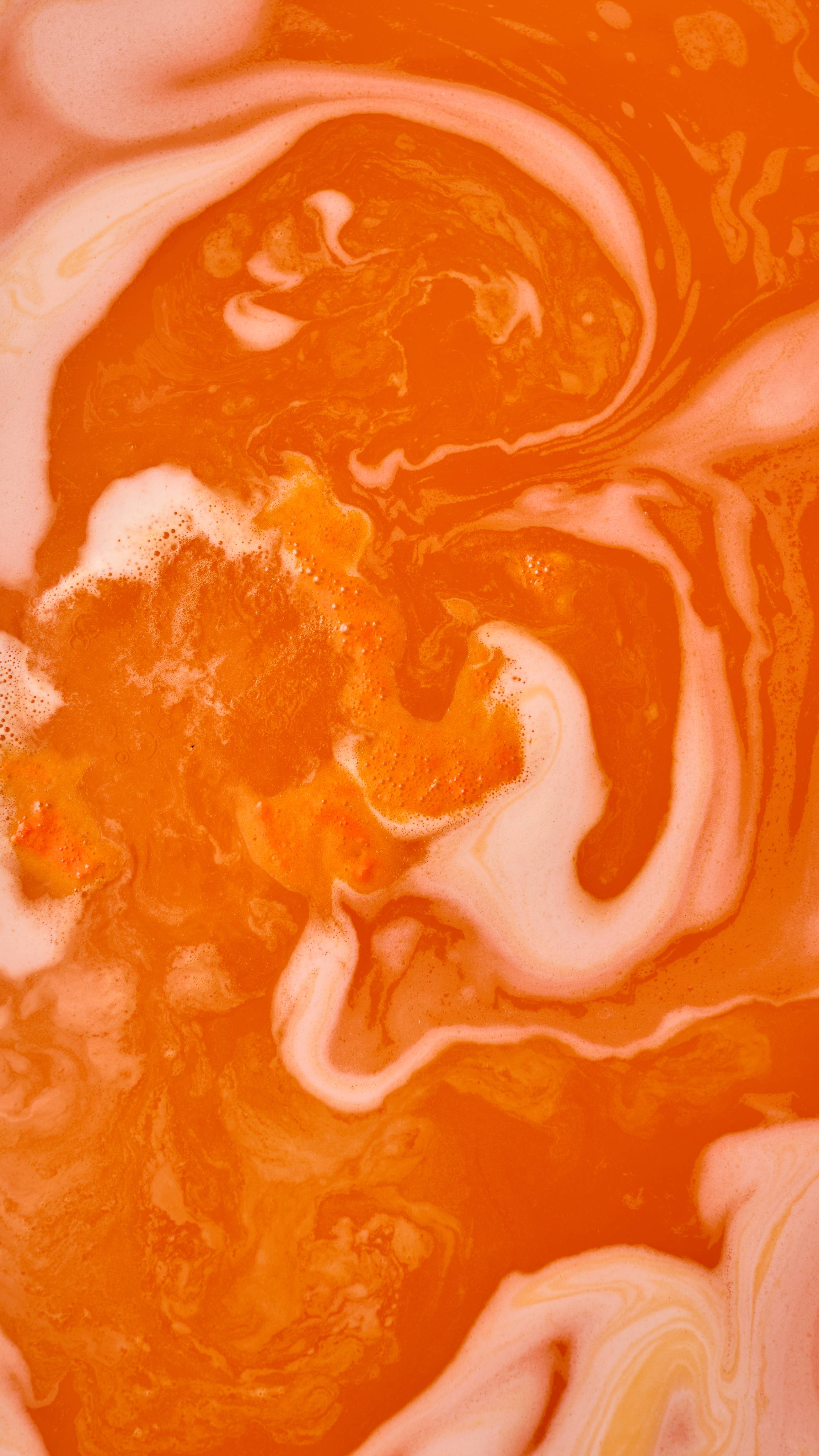 An abstract aerial shot of bath bomb foam swirling in water, creating marbled, wavy patterns of vibrant orange and pale peach-white froth. Clusters of fine, orange-tinted bubbles are scattered across the textured surface.