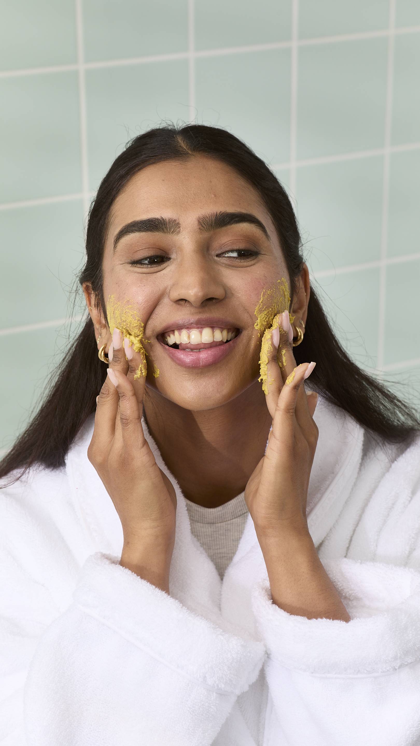 The image shows a model smiling, wearing a white robe, applying a yellow, grainy "Thud Of The Dozy Bumblebee" face cleanser. They are using their fingertips to massage the product onto their cheeks against a backdrop of light green bathroom tiles.