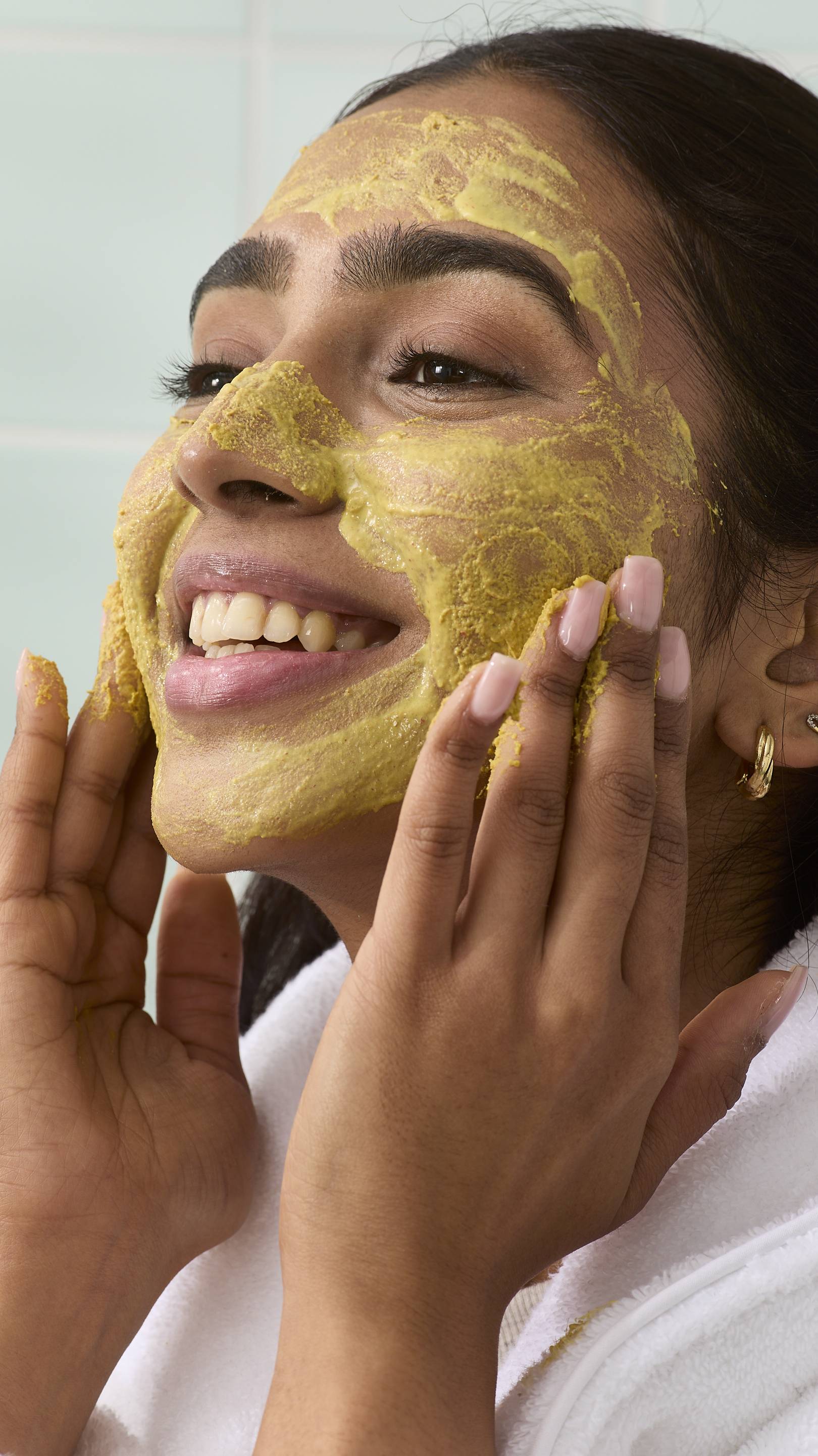 A close-up, side profile of a smiling model wearing a white robe, applying a yellow "Thud Of The Dozy Bumblebee" face cleanser. Their hands massage the product onto their cheeks and forehead against a backdrop of light green bathroom tiles.