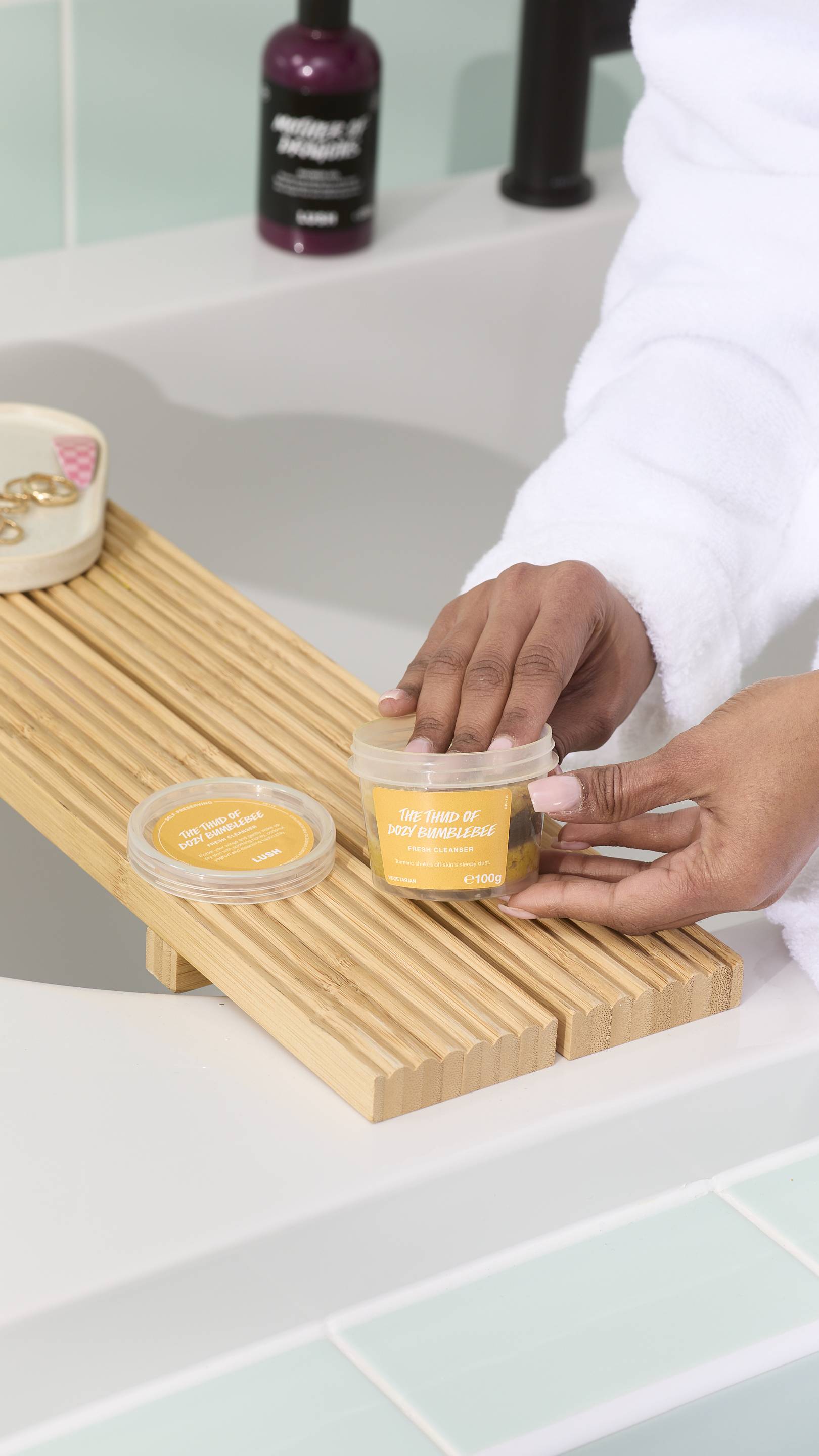 The image shows a model wearing a white dressing gown reaching for a tub of "The Thud of Dozy Bumblebee" fresh cleanser. The product sits on a wooden bath caddy alongside its lid and some gold jewellery, with light green tiles in the background.