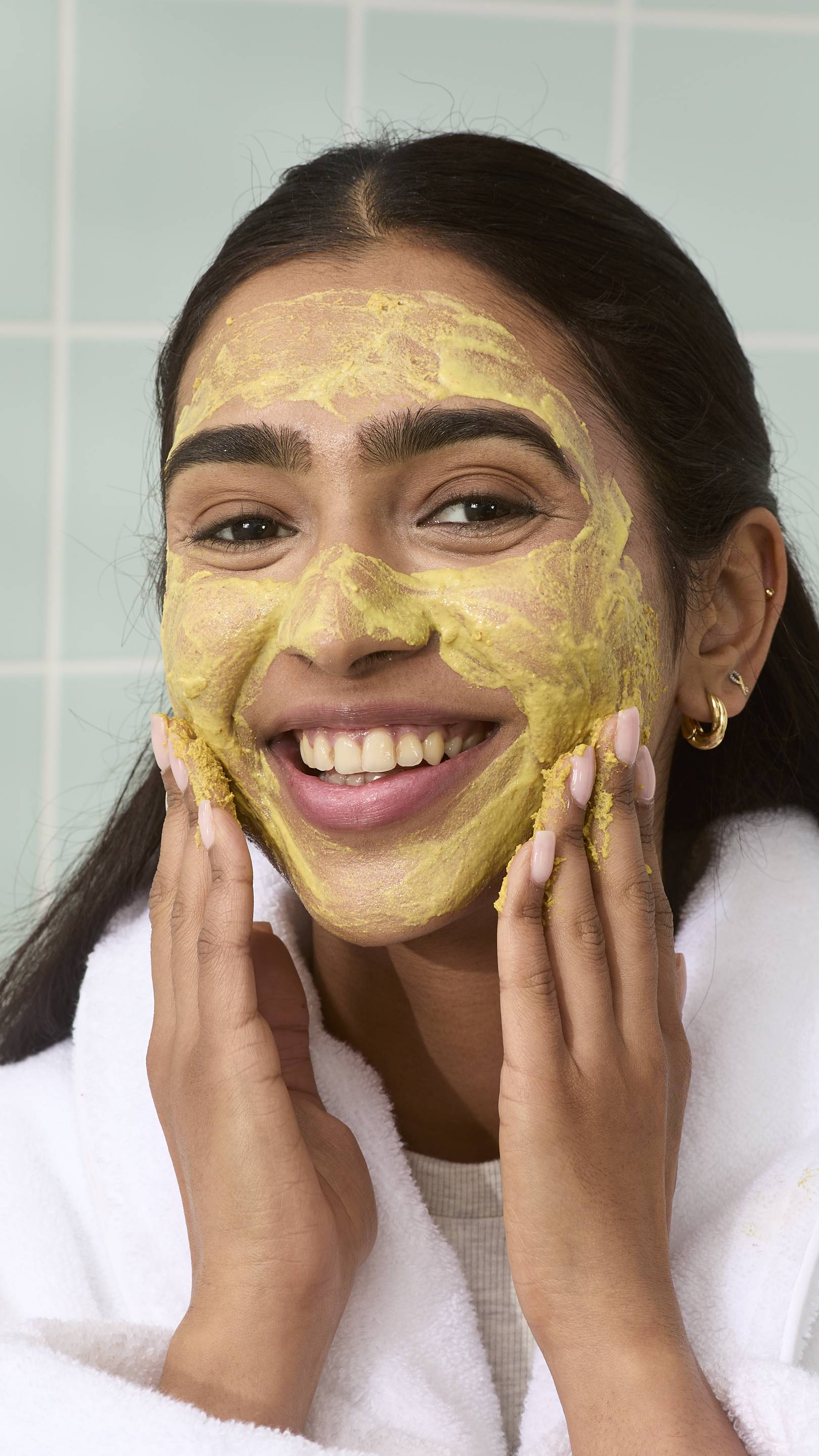 A close-up of a model with long dark hair, wearing a white robe, with a yellow grainy "Thud Of The Dozy Bumblebee" face cleanser spread across most of their face. They are smiling broadly against a backdrop of light green tiles.
