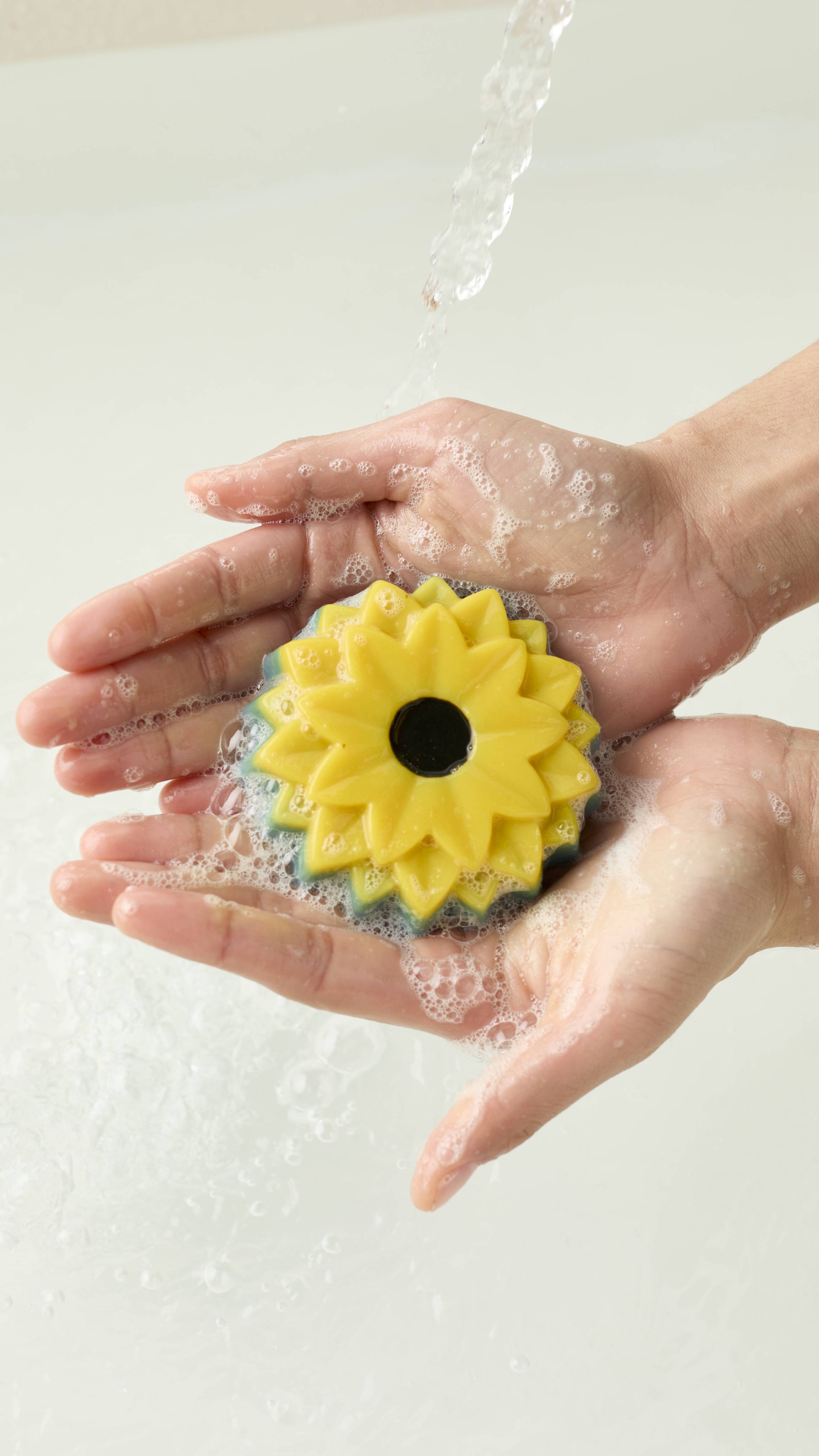 An aerial view of two hands holding the sunflower-shaped There Will Be Sun soap. Water runs over the hands as they create a soapy lather with the product.