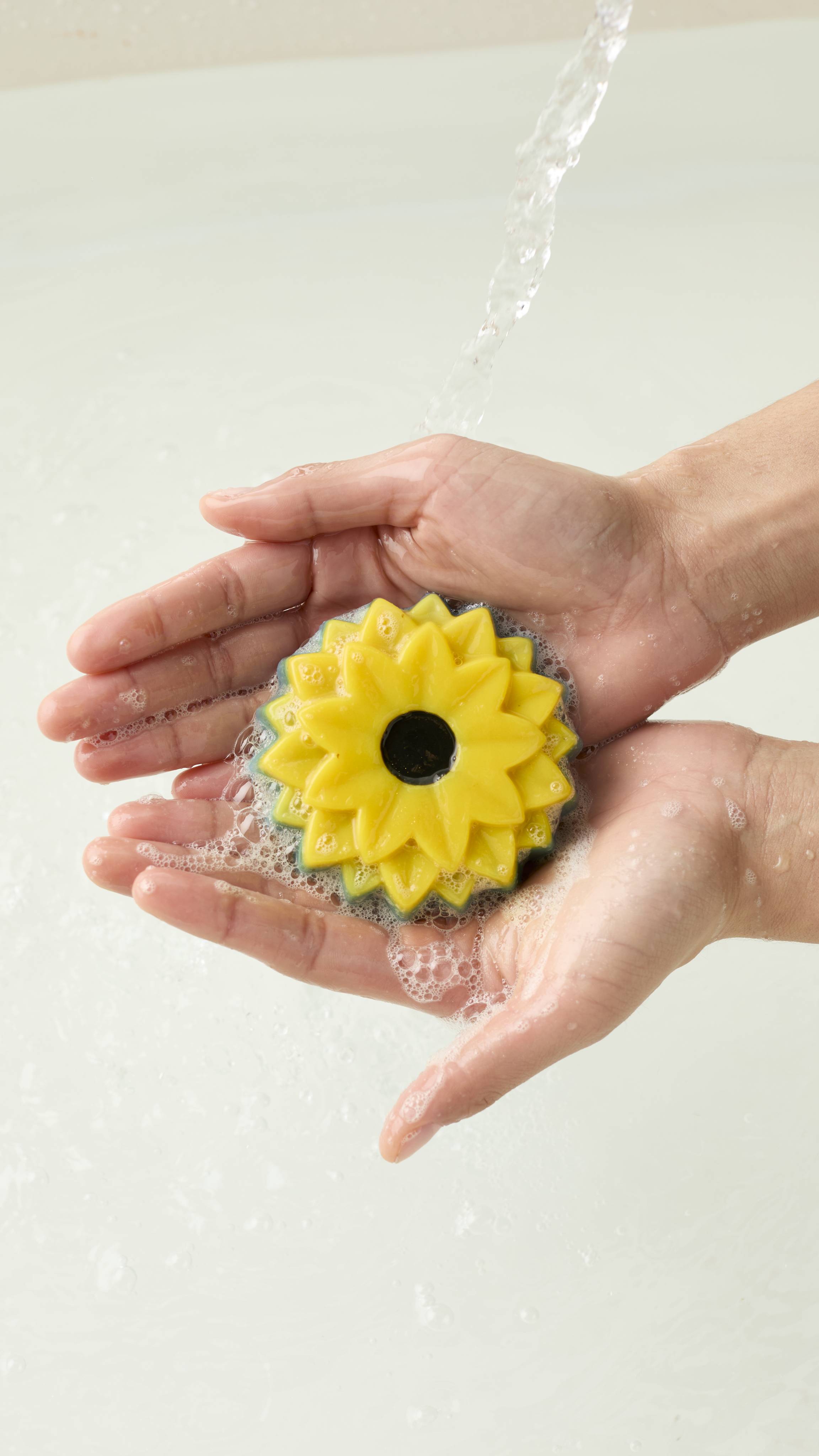 An aerial view of two hands holding the sunflower-shaped There Will Be Sun soap. Water runs over the hands as they create a soapy lather with the product.