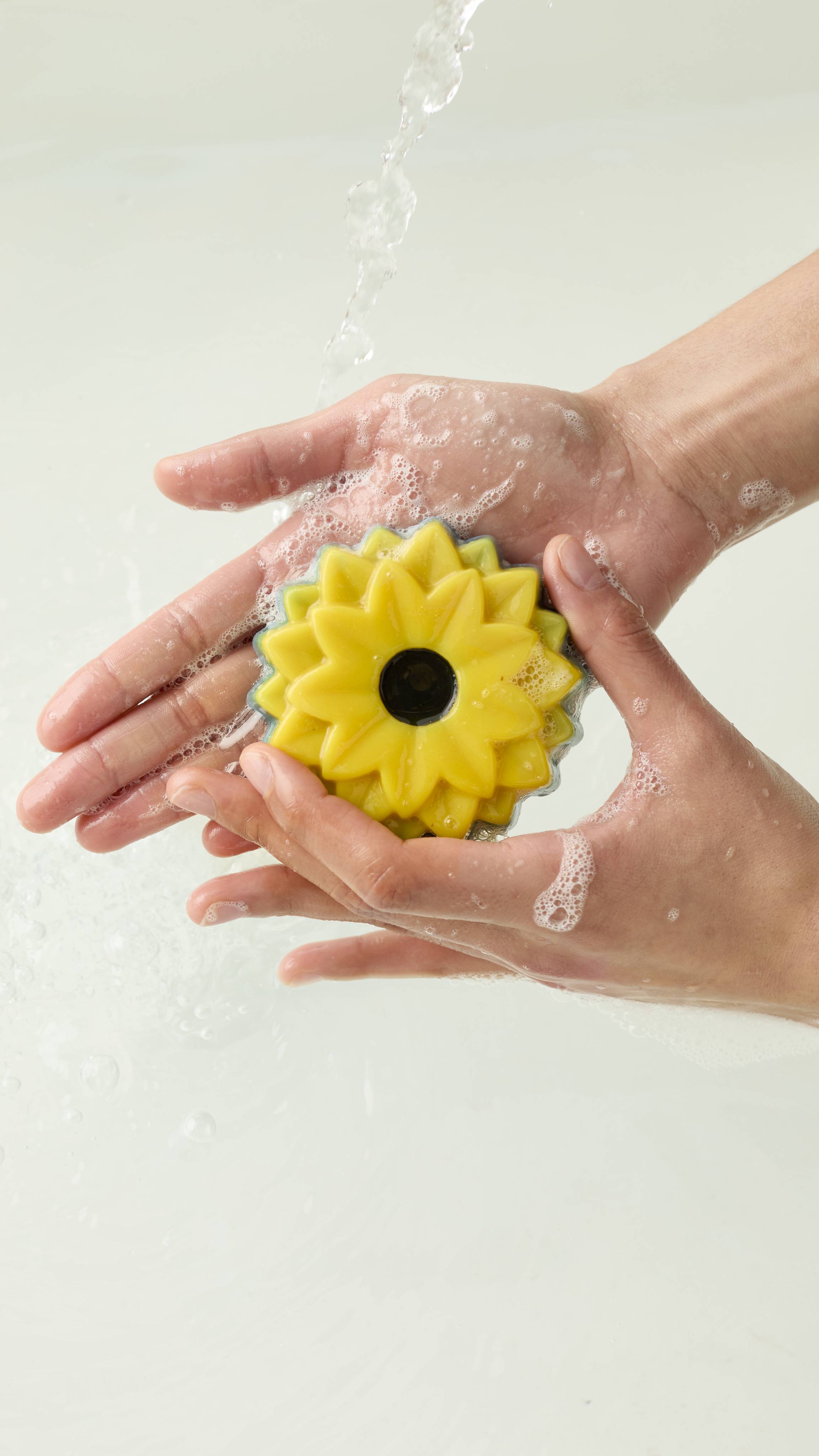 An aerial view of two hands holding the sunflower-shaped There Will Be Sun soap. Water runs over the hands as they create a soapy lather with the product.